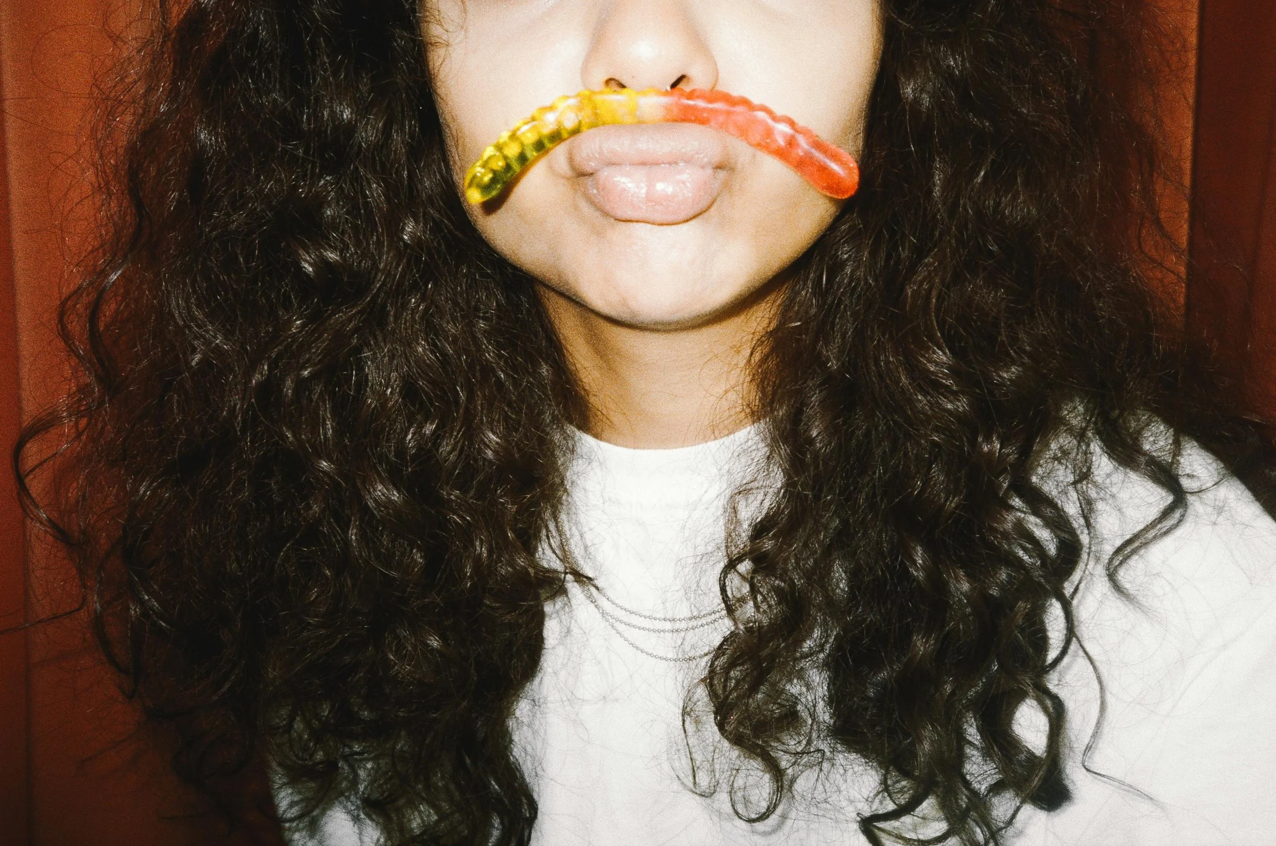 Person with curly dark hair holding gummy worms in mouth, forming a mustache.