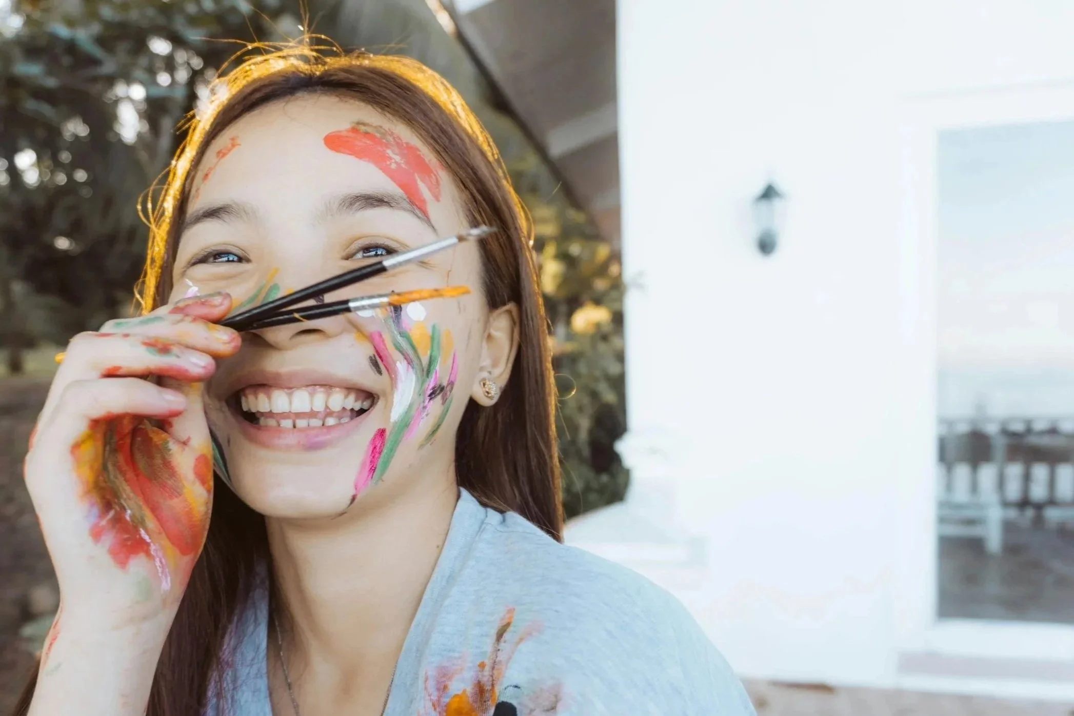 Young woman smiling with face and hand covered in colorful paint, holding paintbrushes near her face outdoors on a sunny day.