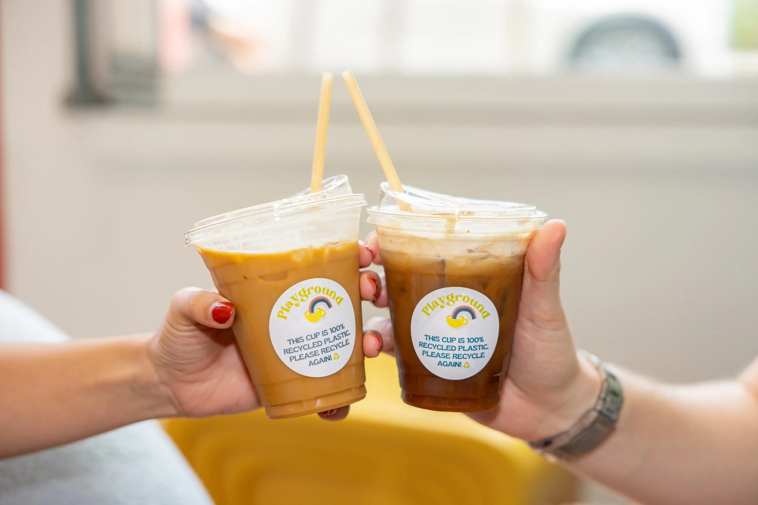 Two people holding plastic cups filled with iced coffee or tea, clinking together in a toast. The cups have a label with a rainbow and bear the message about recycling plastic.
