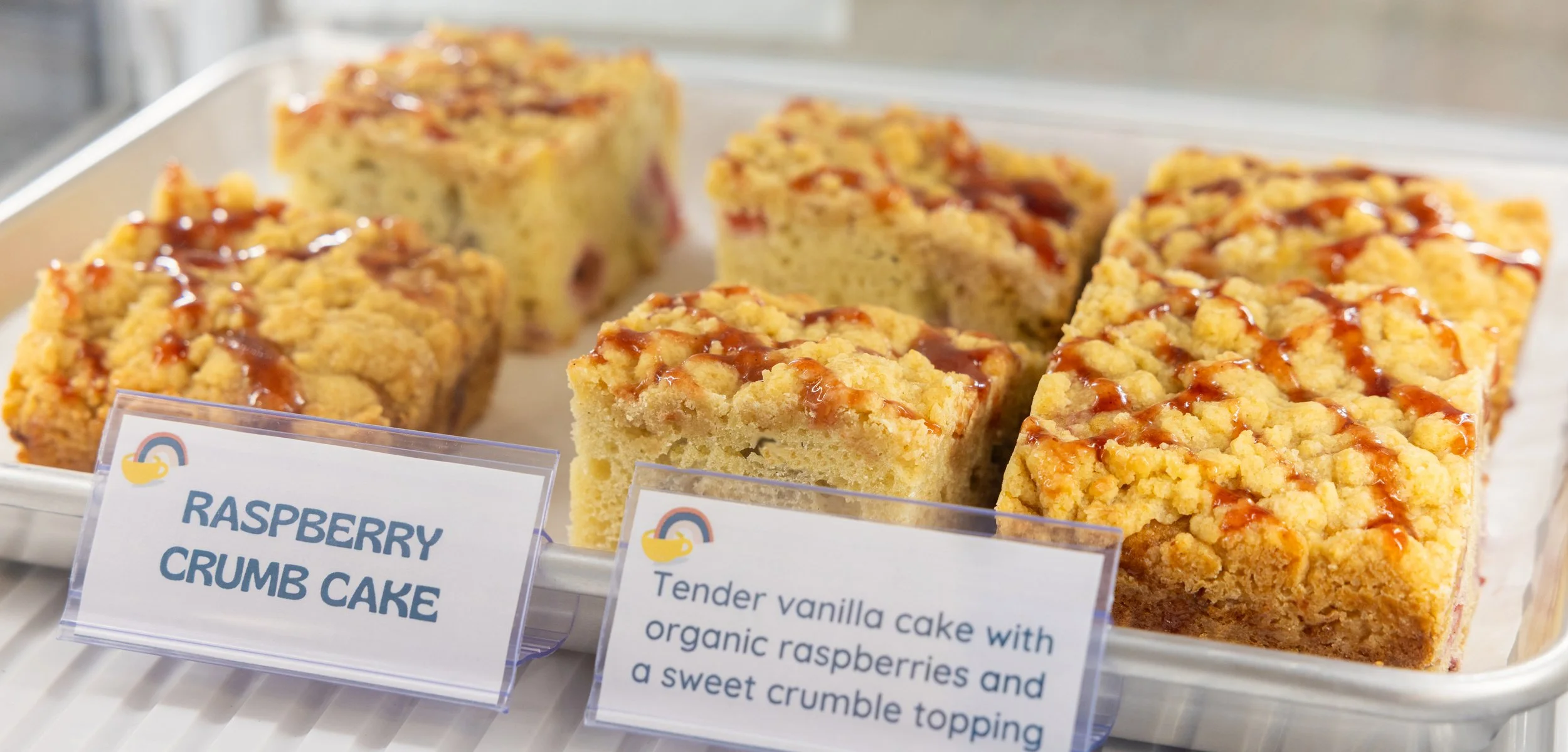 Close-up of raspberry crumb cake squares with crumb topping and drizzled caramel, displayed in a bakery case with labeled signs.