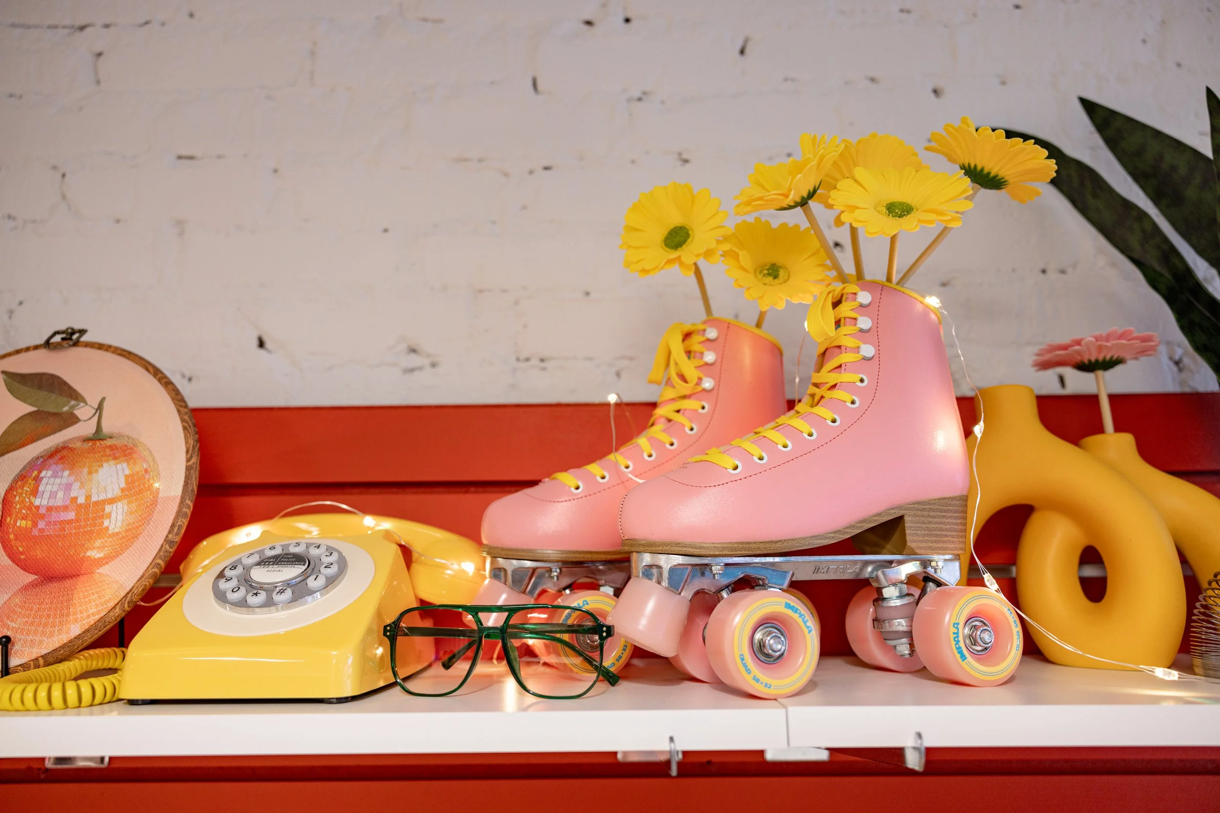 Pink roller skates with yellow laces and flowers growing out of the top, placed on a white shelf along with vintage yellow rotary phone, a pair of green glasses, and other colorful decor items against a white brick wall.