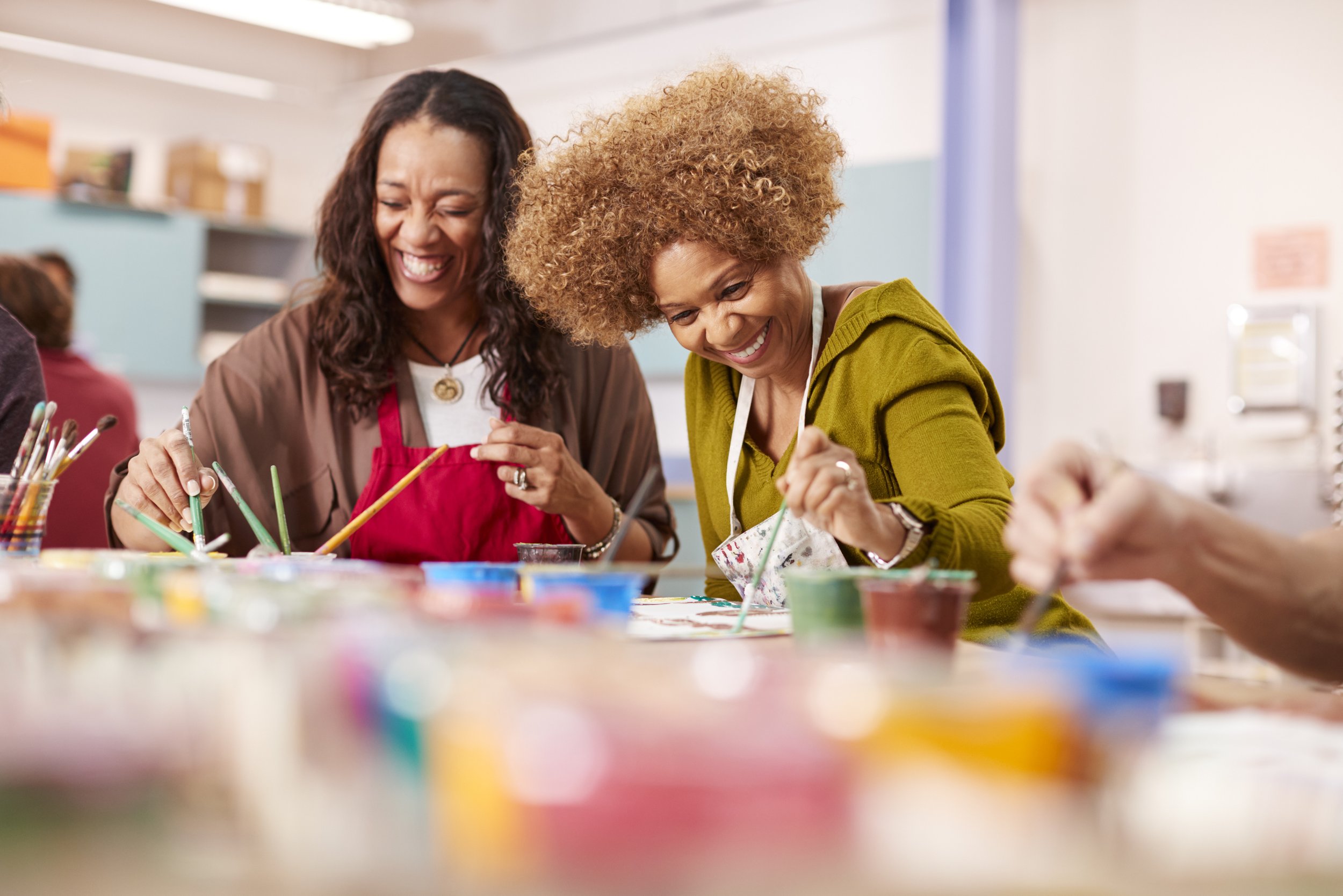 Two women painting at a table with colorful paints, smiling and enjoying an art activity together.