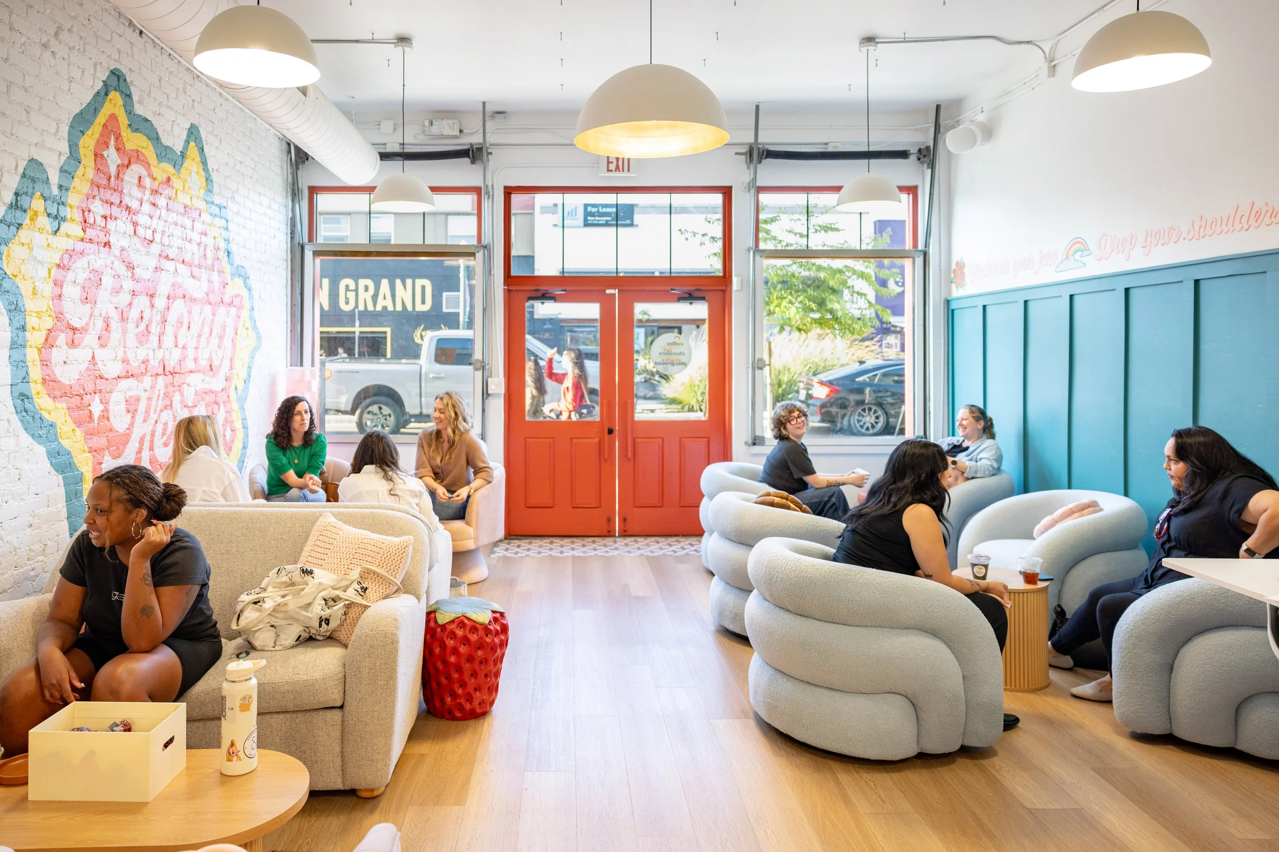 A cozy, brightly lit coffee shop interior with white brick walls, colorful wall art, and several people sitting on plush chairs and couches, engaging in conversations. Large windows and glass doors provide natural light, with some patrons visible outside. Overhead pendant lights hang from the ceiling.
