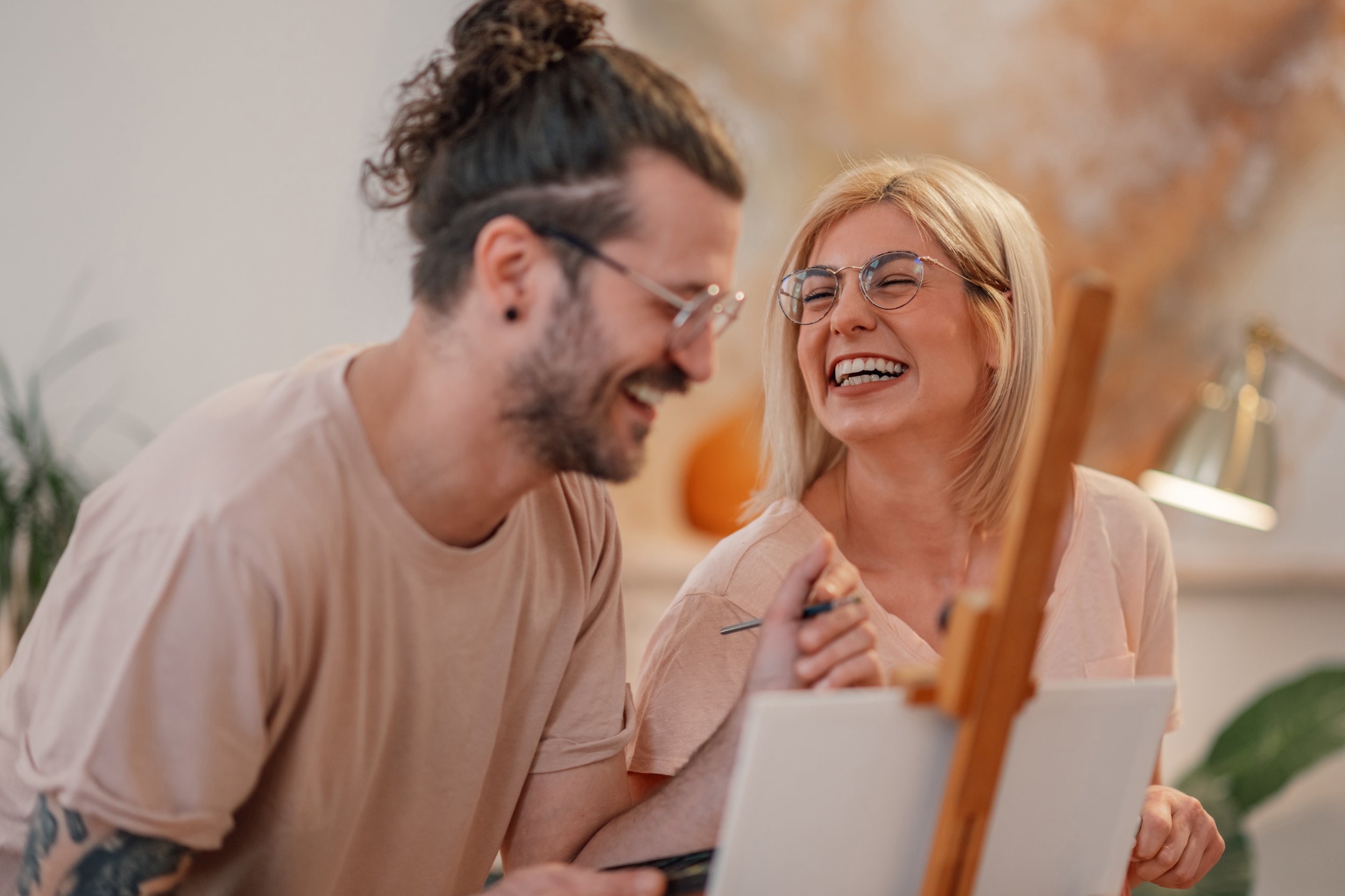 Two people, a woman and a man, laughing and painting together in a cozy room. The woman is holding a palette and brush, and both are wearing glasses.