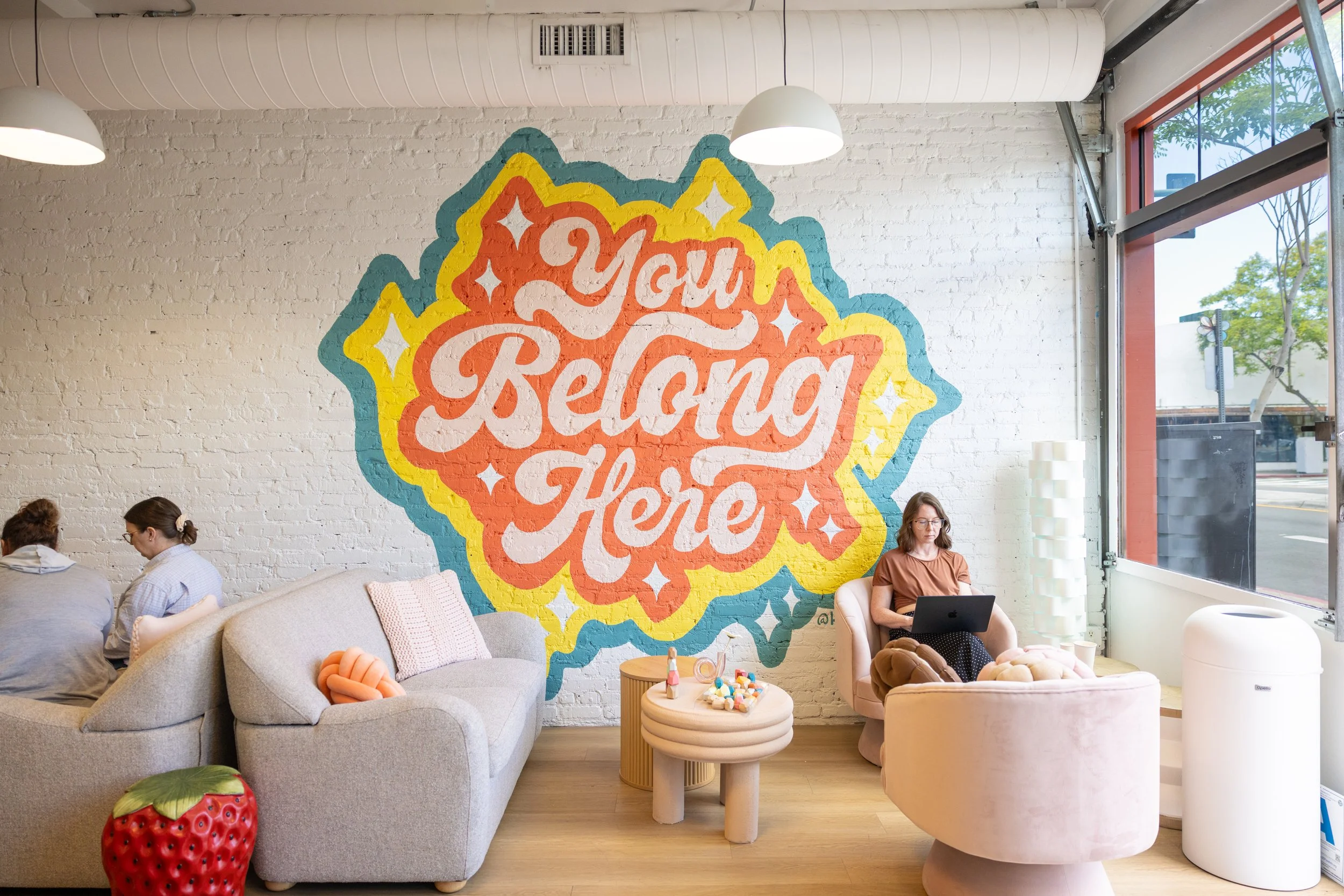 Interior of a cafe with white brick walls, a colorful mural with the words "You Are Here," and large windows showing trees outside. There are several people sitting on couches and chairs, some using laptops, and a small table with decorations and books.