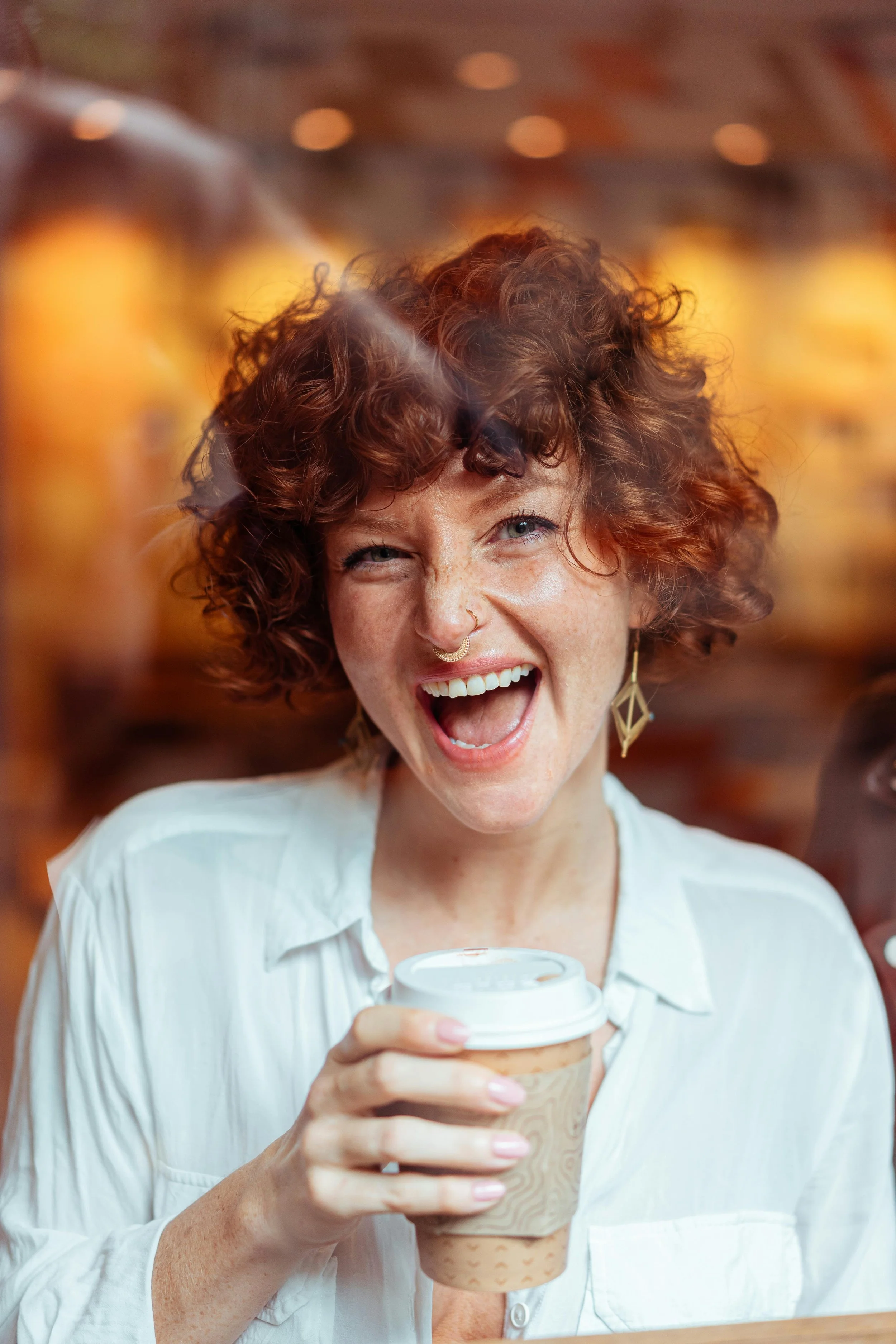 A woman with curly red hair and a nose ring is smiling and laughing, holding a to-go coffee cup in a coffee shop.