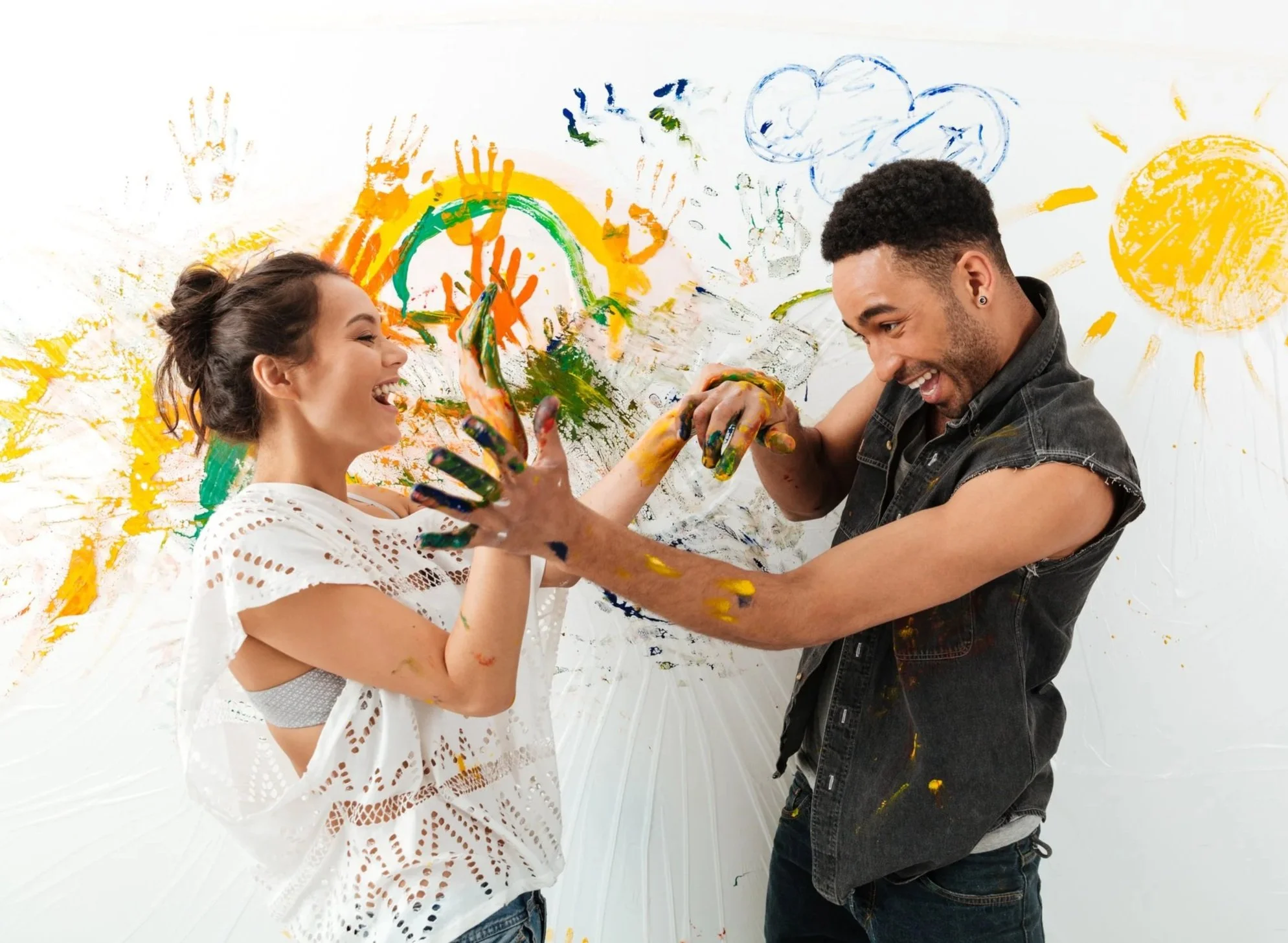 A woman and a man playing with paint against a wall with colorful handprints, rainbows, clouds, and a sun.