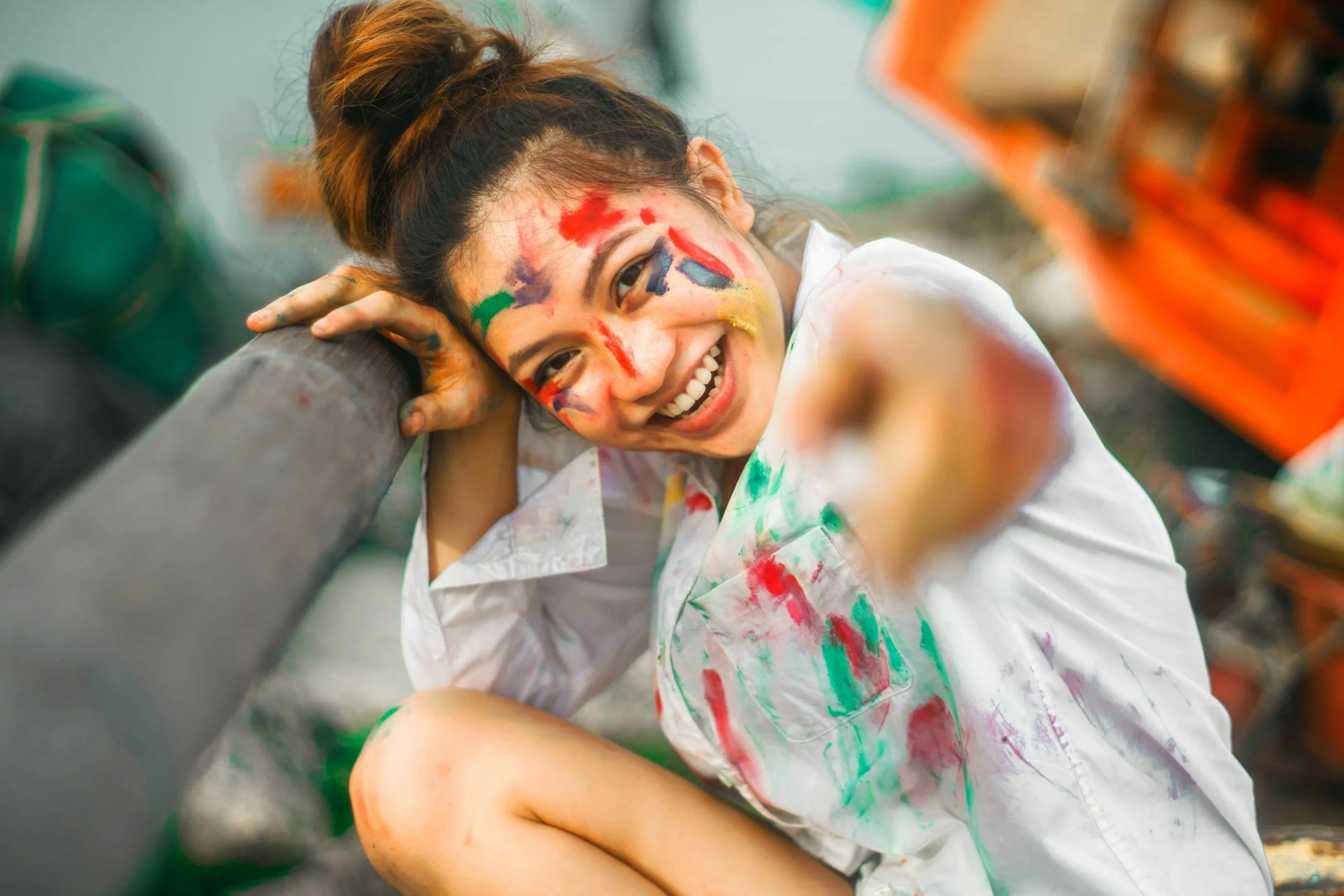 A young woman with a bright smile and colorful face paint, wearing a white shirt with paint stains, leaning on a pipe or pole, with paint splatters on her face and shirt.