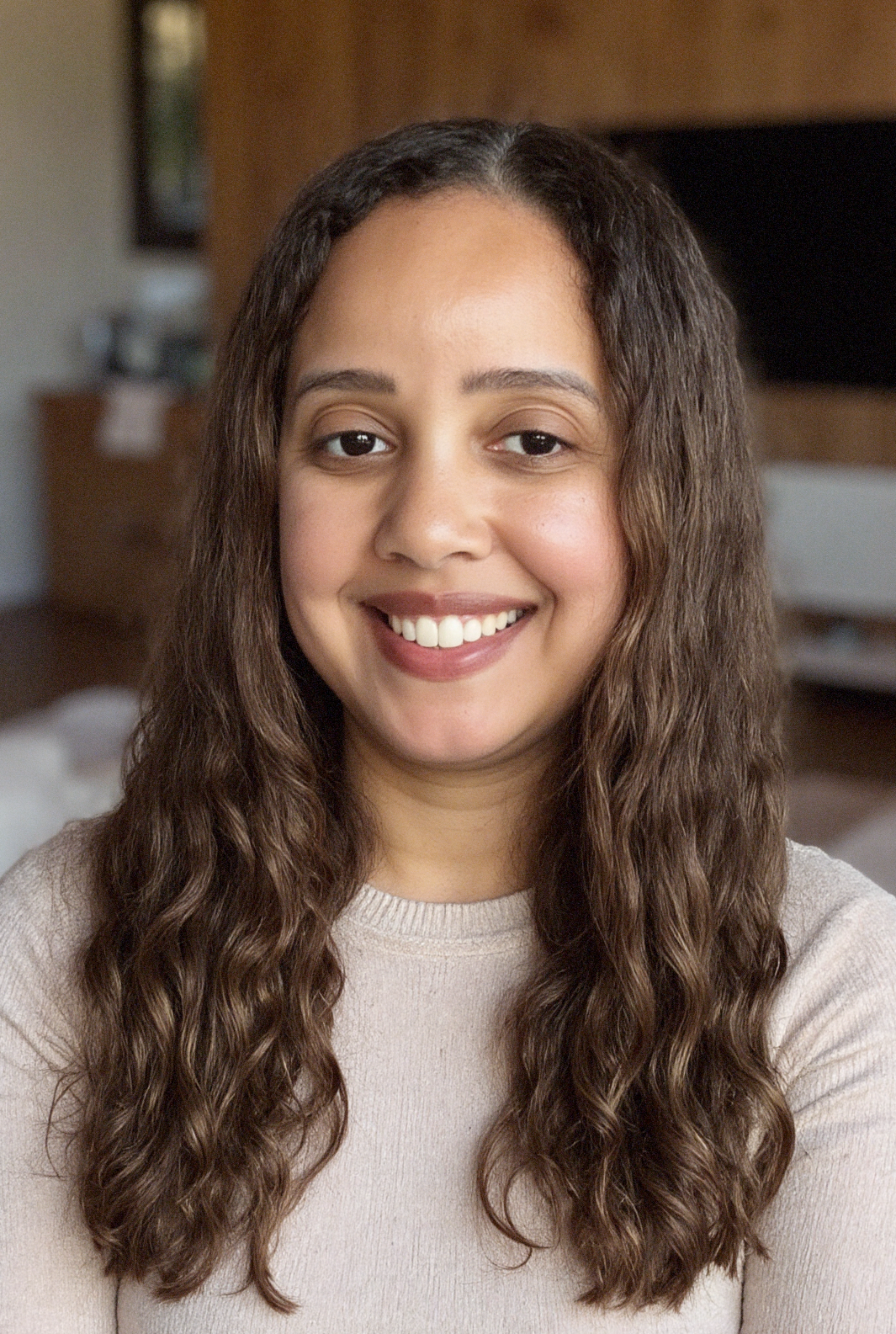 A woman with long, wavy brown hair, smiling, wearing a beige sweater, in a cozy interior with a wooden wall and a television in the background.