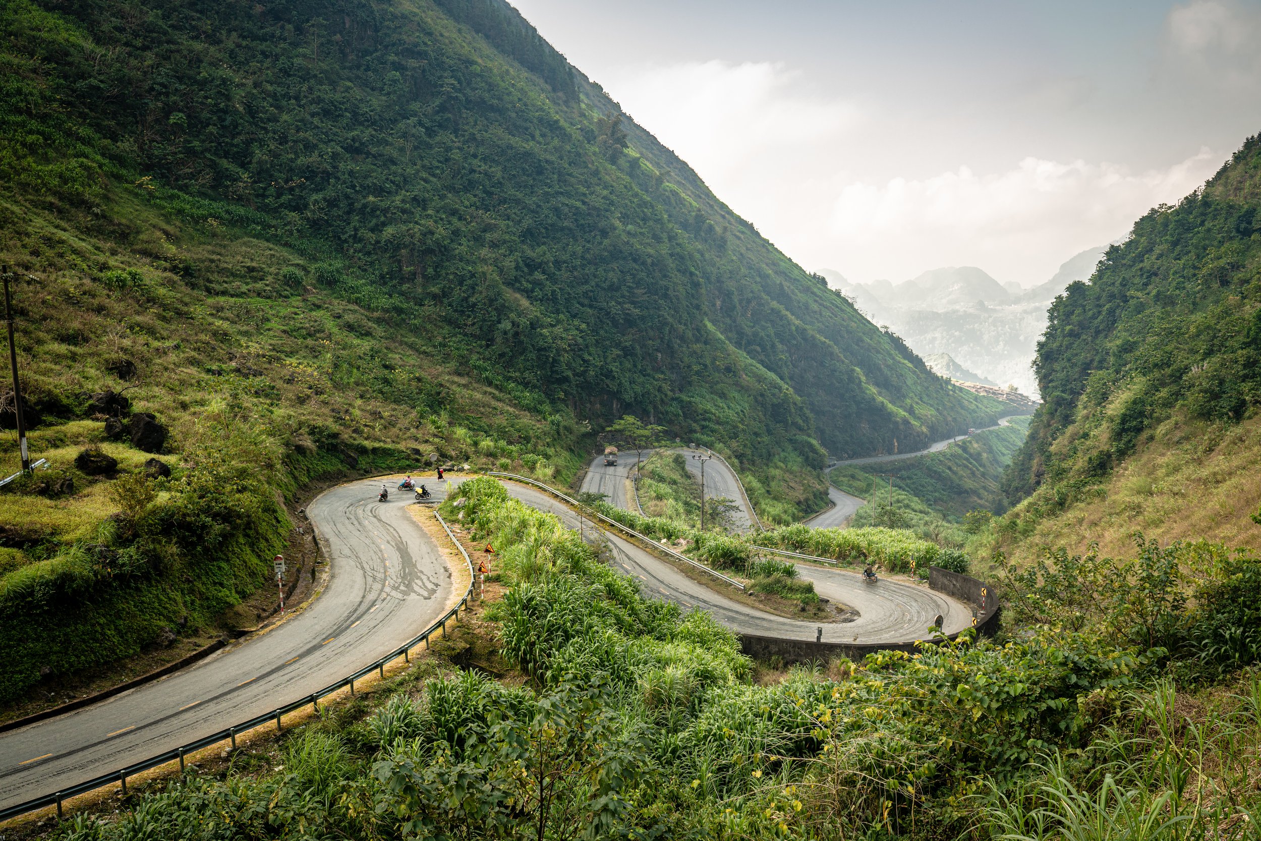 A winding mountain road in a lush green valley with tall hills on both sides, some vehicles and motorcycles traveling along the road, and a cloudy sky overhead.