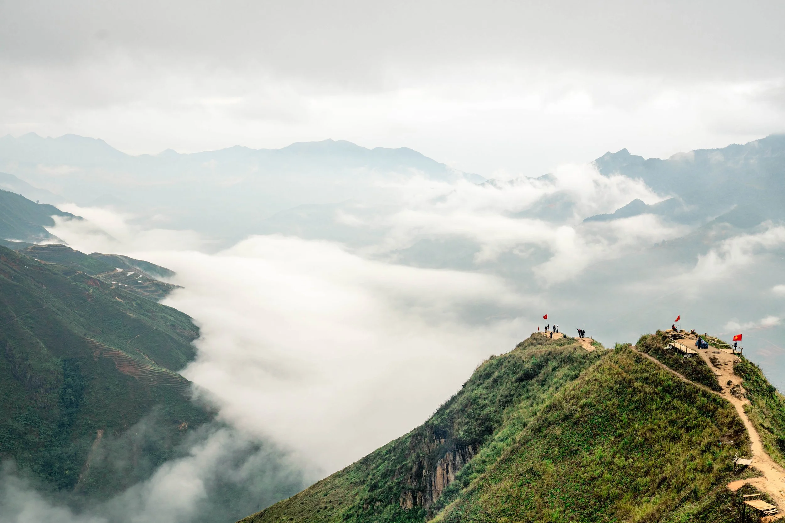 Mountaintop with small flags, people, and dogs on a dirt trail surrounded by clouds and green hills.