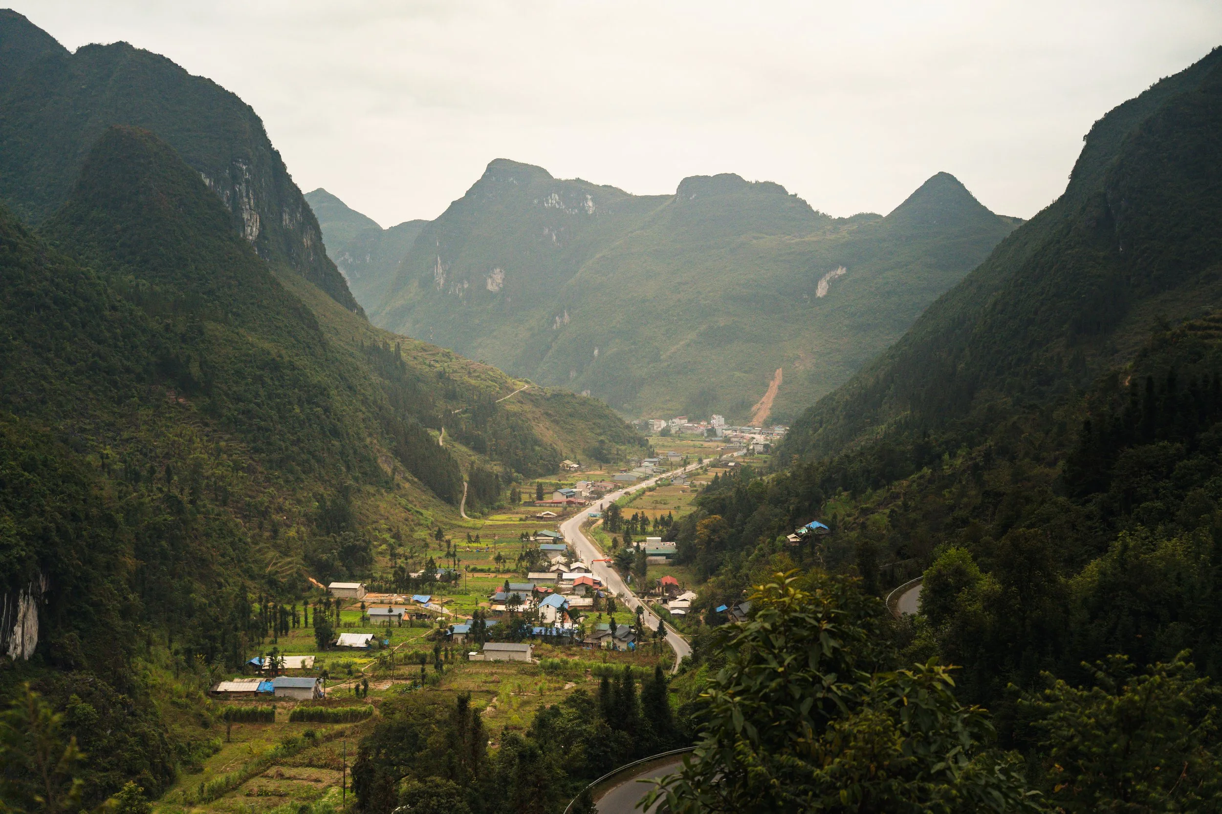 A small village in a lush green valley surrounded by tall, steep mountains with winding roads and farmland.