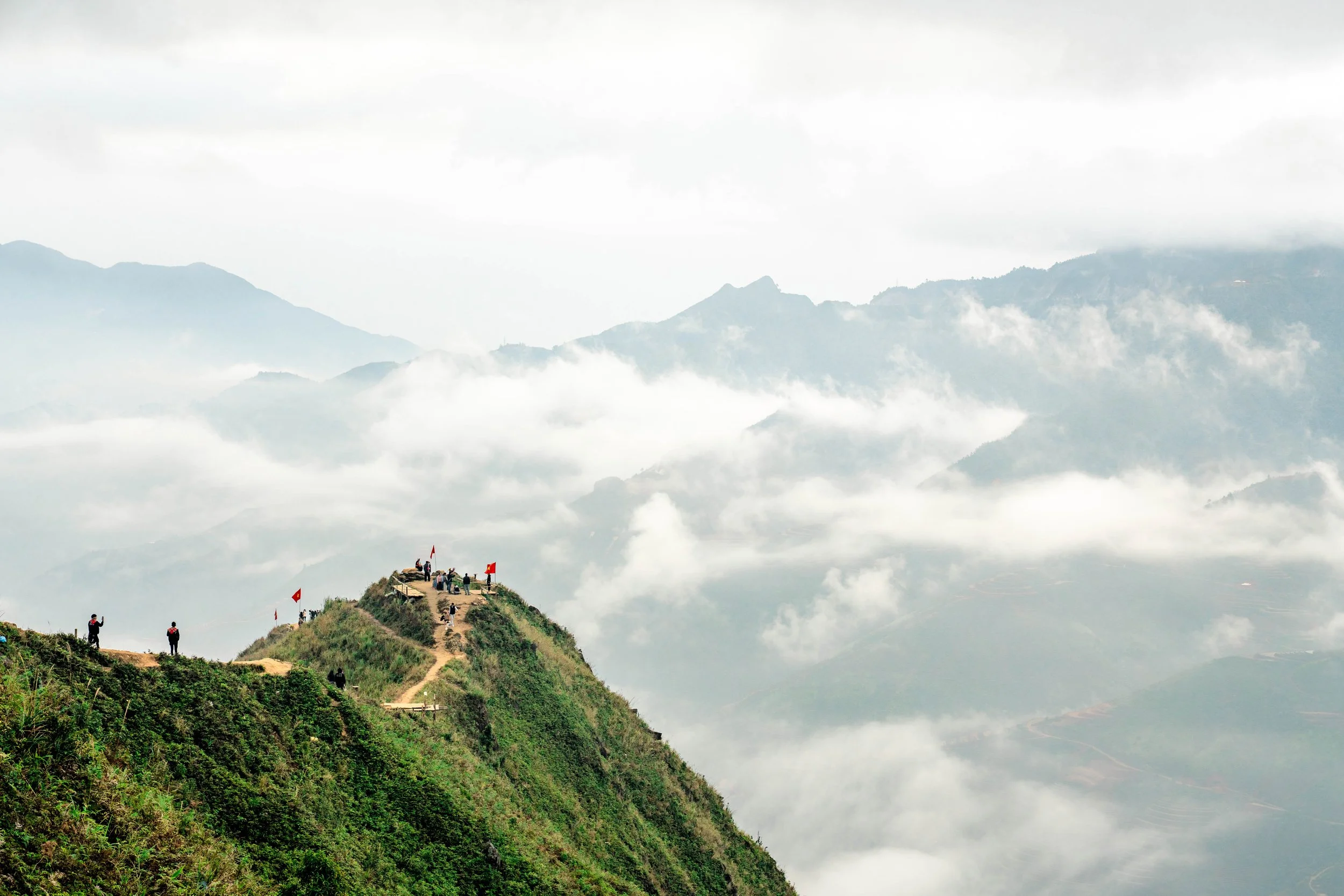 A mountain ridge with a trail and several people, some taking photos and others standing or sitting, with red flags on the ridge, overlooking cloud-covered mountains in the distance.