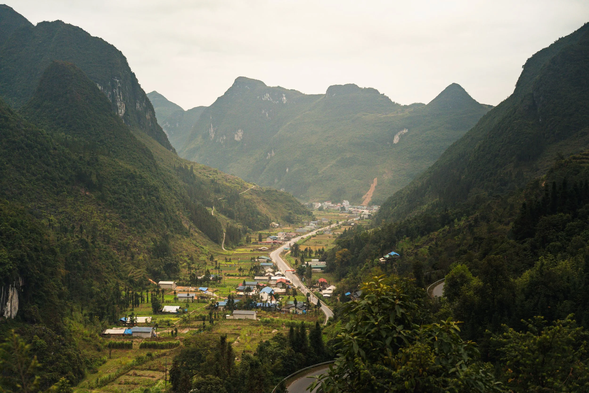 A scenic view of a mountain valley with a winding road, small houses, and lush greenery, surrounded by tall mountains under a cloudy sky.