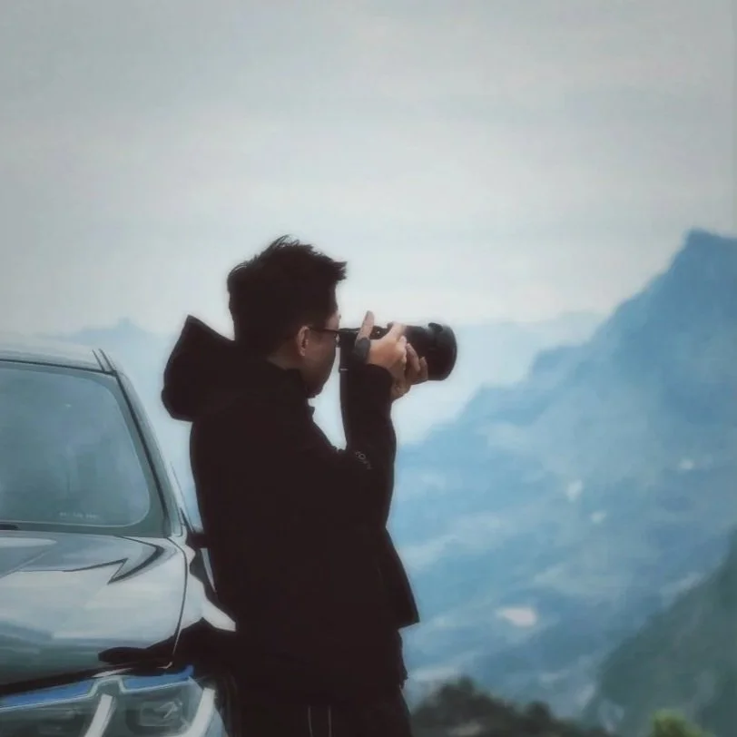 A person taking a photograph outdoors, standing next to a car, with mountains and cloudy sky in the background.