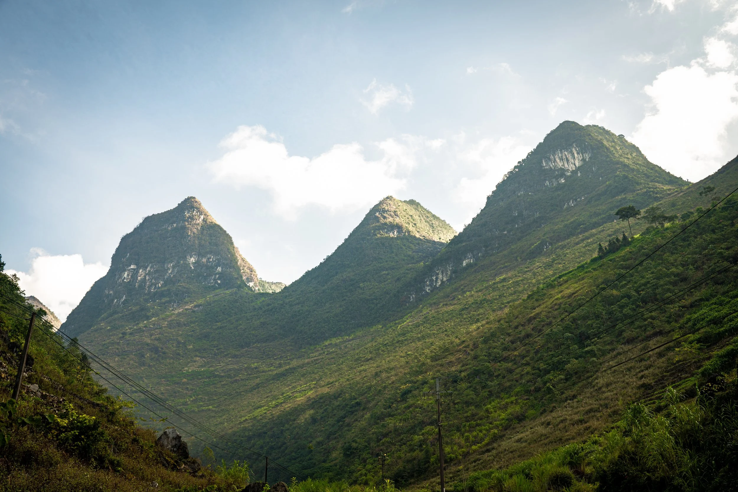 Green mountains with rugged peaks under a partly cloudy sky, with power lines in the foreground.