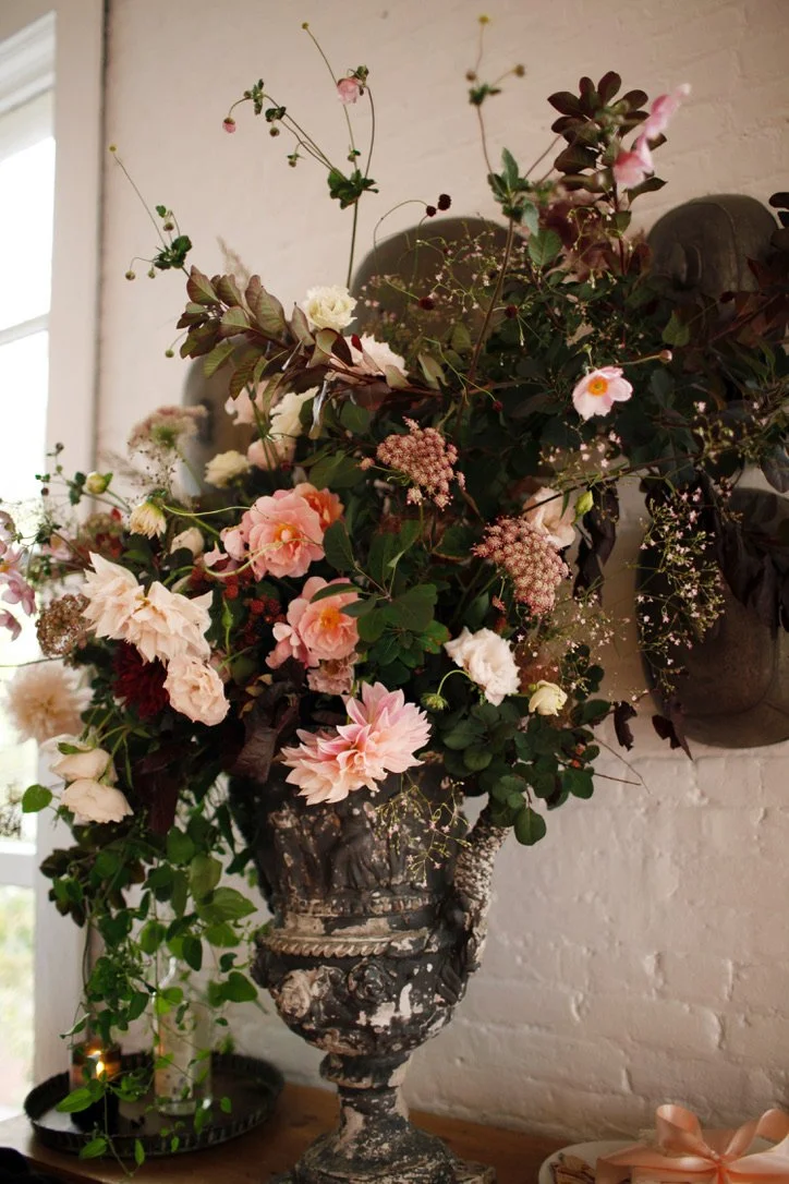 A large, ornate metal vase filled with a variety of pink, white, and burgundy flowers and greenery, placed on a wooden surface against a white brick wall.