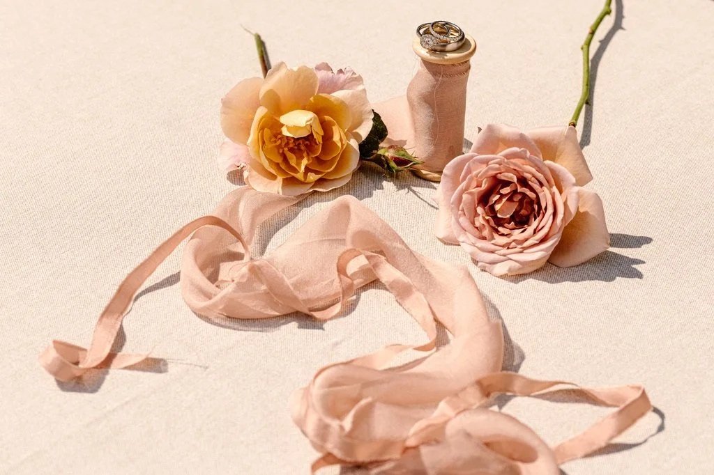 Pink roses, a spool of pink ribbon, and wedding rings on a light-colored fabric background.