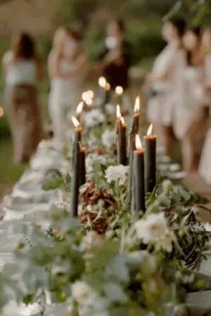 Candles and floral arrangements on a long table at an outdoor gathering, with blurred people in the background.