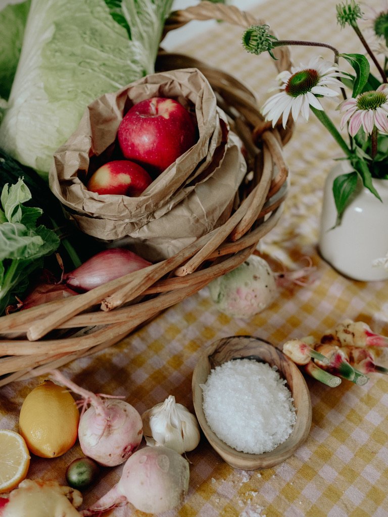 Fresh apples in paper wrapping in a wicker basket, along with assorted vegetables, garlic, and salt on a yellow checkered tablecloth, with a white vase of flowers nearby.