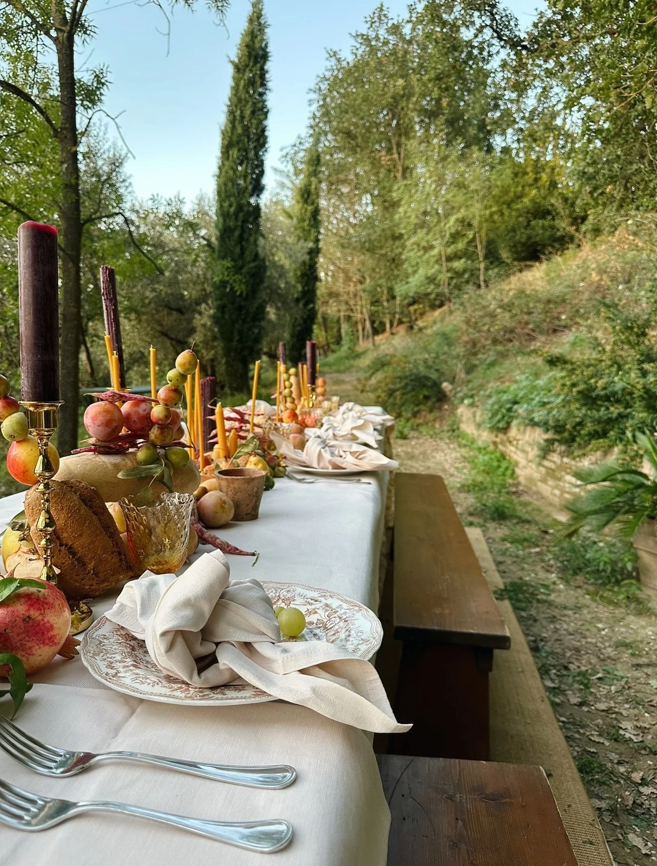 An outdoor dining table set against a wooded background, decorated with a white tablecloth, fruit, candles, and tableware.