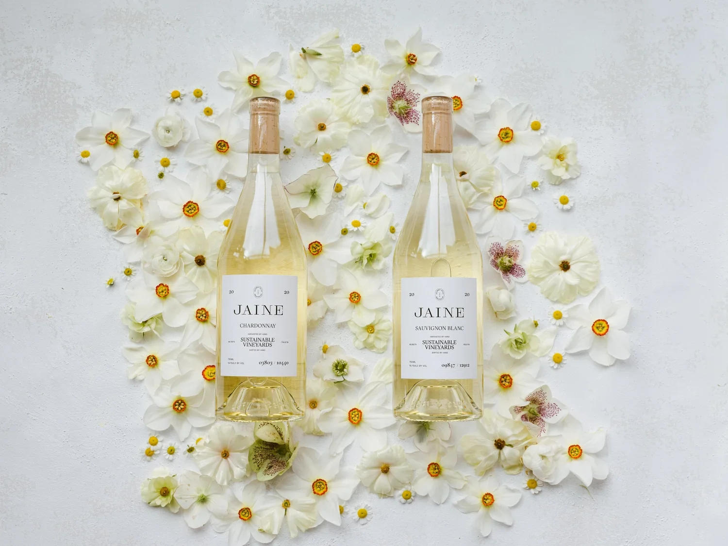 Two bottles of Jaine wine, Chardonnay and Sauvignon Blanc, placed on a white surface surrounded by white and pink flowers.