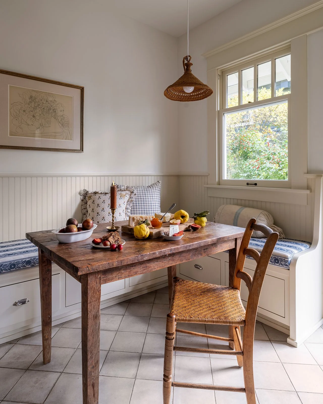 A cozy kitchen nook with a wooden table set for breakfast, featuring a bowl of apples, a loaf of bread, and yellow and green apples. There are pillows on the built-in corner bench, a framed picture on the wall, and a window letting in natural light with a view of greenery outside. A wicker pendant lamp hangs from the ceiling.