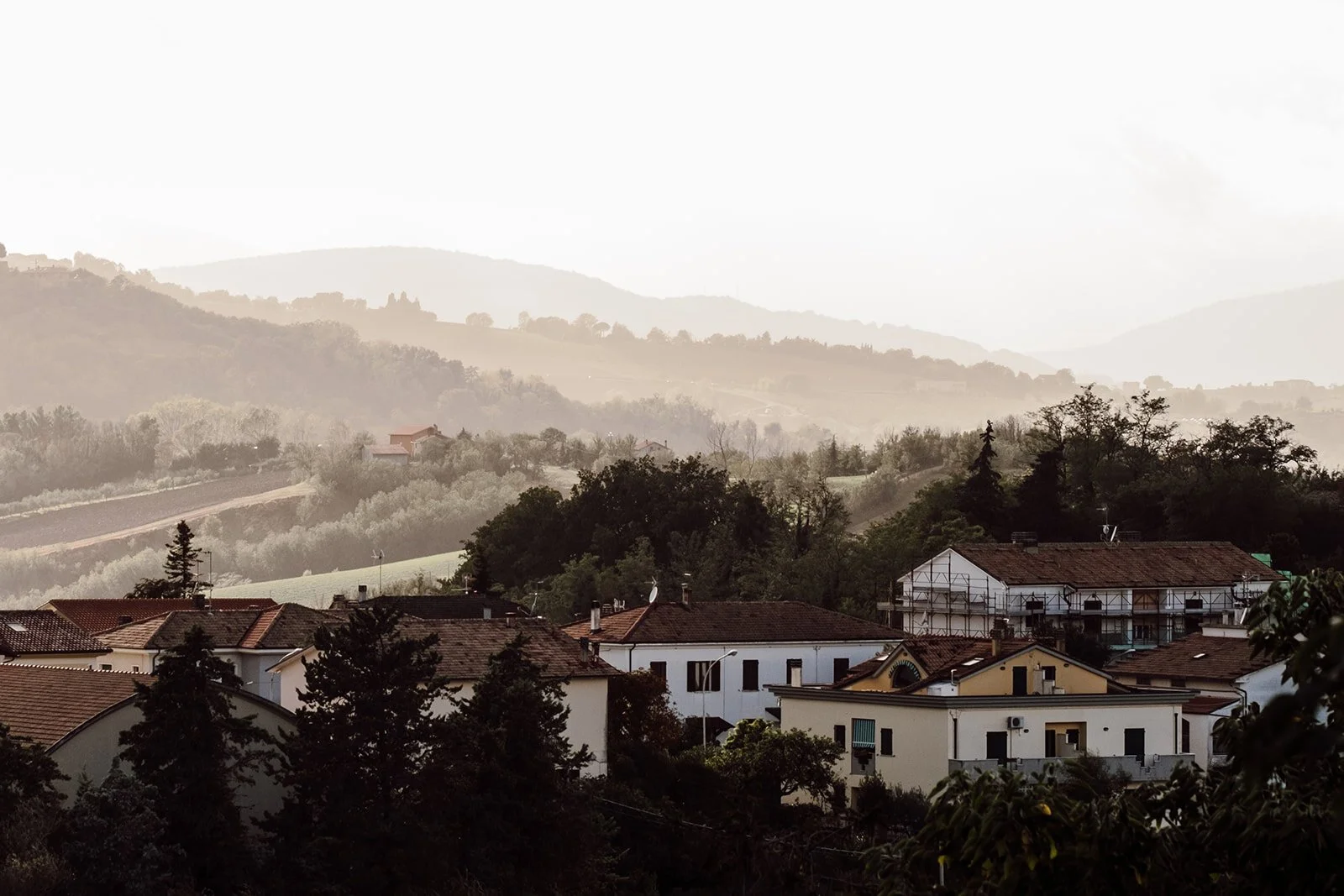 Hilly landscape with houses in the foreground and layered hills in the background, some with trees and fields, under a cloudy sky.
