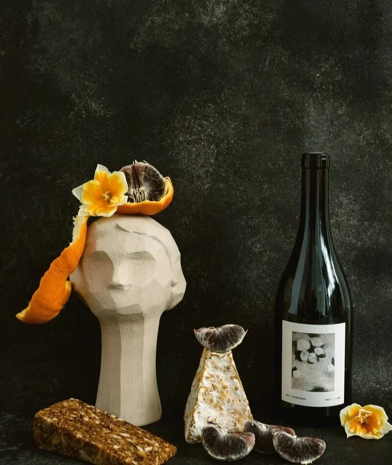 A still life composition with a wooden faceless bust, an orange peel, a lemon flower, a rock, a mushroom, and a bottle of wine against a dark background.