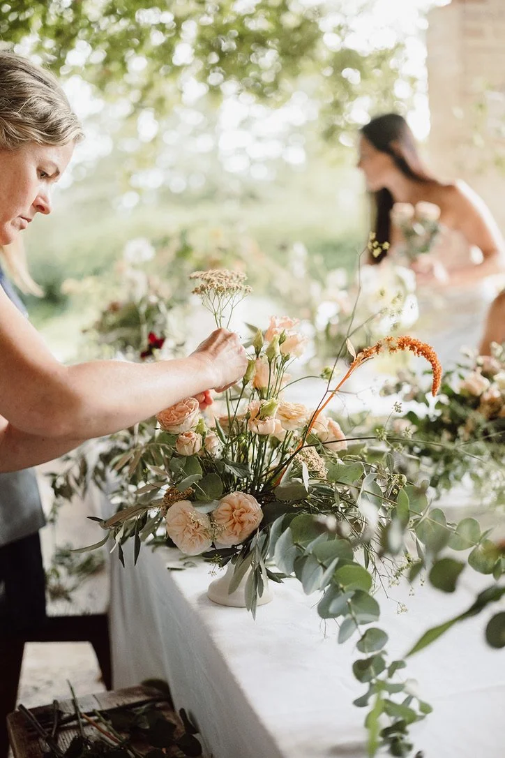 A woman arranging a floral centerpiece with peach roses, greenery, and white flowers on a table outdoors, with another woman in the background holding flowers.