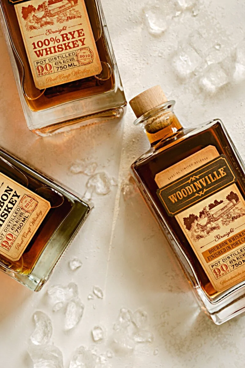 Three bottles of whiskey on a white surface with ice cubes scattered around.