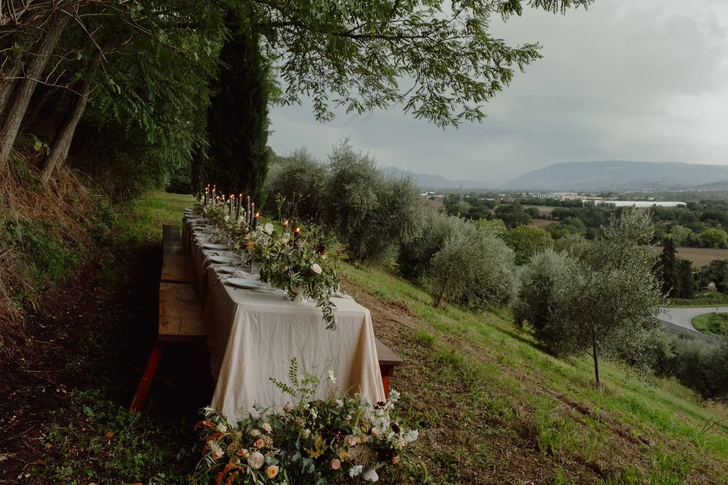 Long outdoor dining table set on a hillside with greenery, trees, and distant mountains under an overcast sky, decorated with candles, flowers, and tableware for a gathering or celebration.