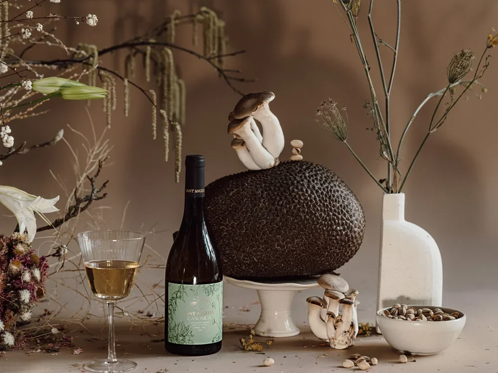 A still life arrangement featuring a bottle of white wine, a glass filled with white wine, various mushrooms including maitake and shiitake, a large textured black mushroom, dried flowers, and a tall white vase with dried grasses and seed pods, all set against a neutral background.