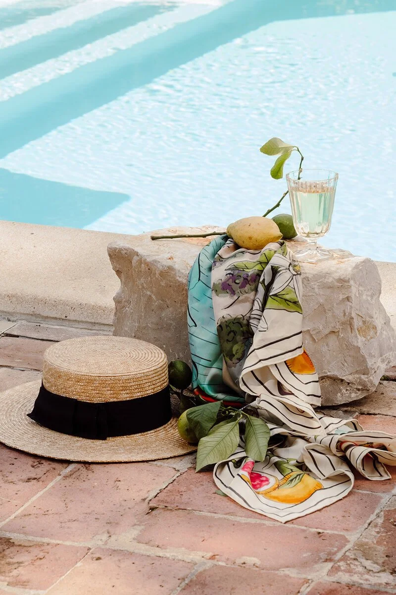 A straw hat with a black ribbon, a glass of water, a scarf with a floral pattern, a lemon, and green fruit on a stone near a swimming pool.