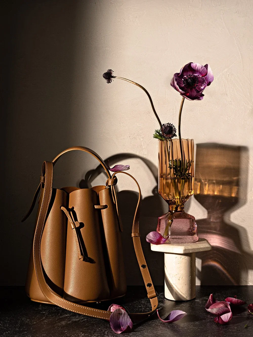 A brown leather handbag with a long strap, a purple flower in a glass vase with pink tint, sitting on a small marble pedestal, with petals scattered around on a dark surface, and shadows cast on the wall behind.