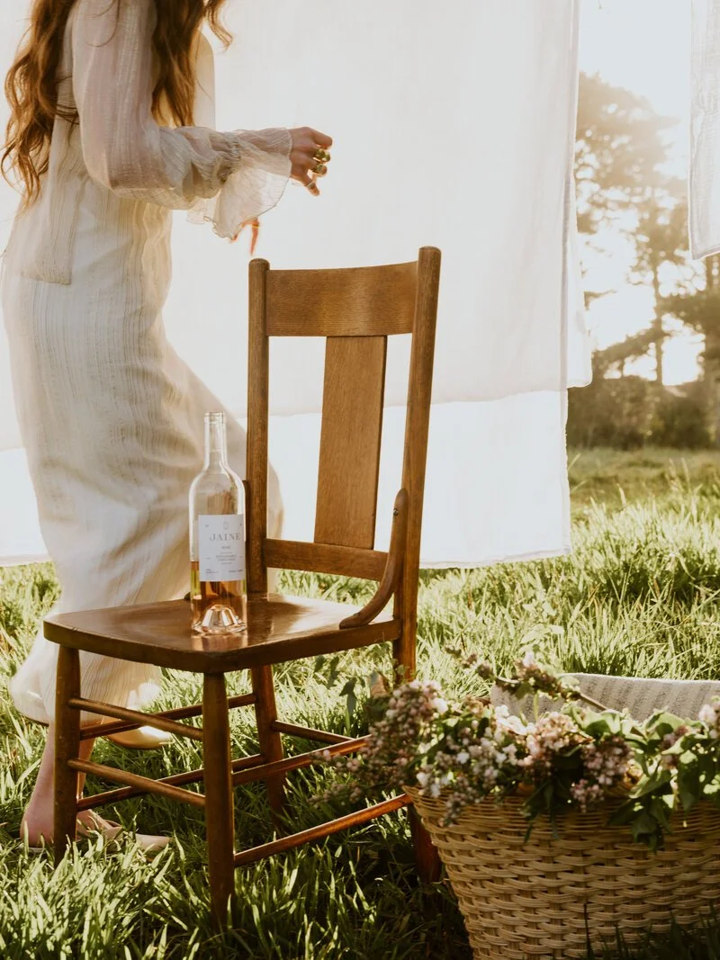 A woman dressed in a light-colored dress standing outdoors next to a wooden chair on green grass, with a bottle of rosé wine on the chair, a basket of flowers nearby, and a white curtain hanging in the background with sunlight shining through.