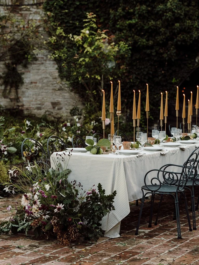 A long outdoor dining table decorated with tall candles, greenery, and flowers, set on a brick patio surrounded by lush greenery and a stone wall.