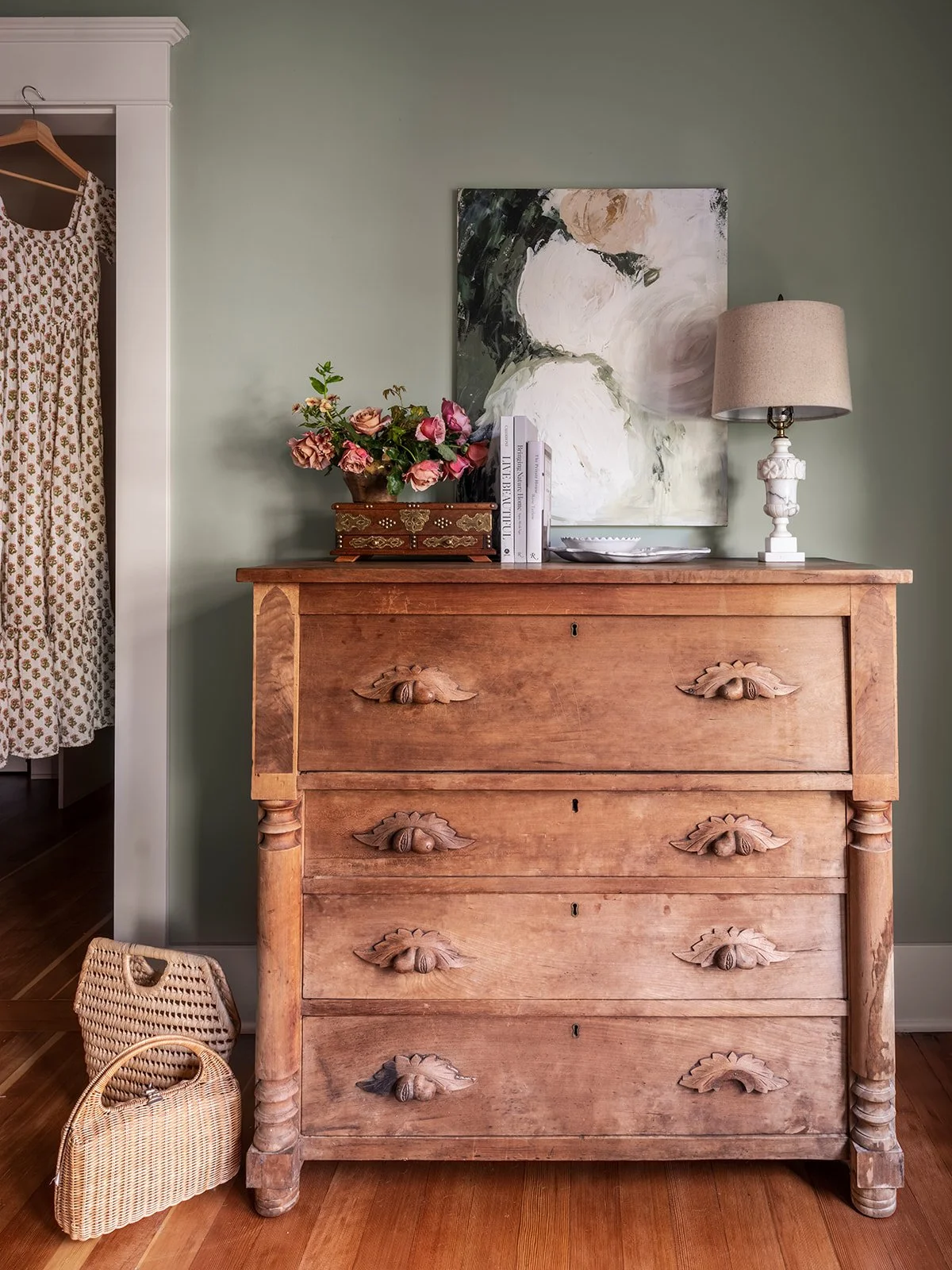 A vintage wooden dresser with flower carvings on the drawer handles, topped with a flower bouquet, books, a decorative box, a white dish, and a table lamp. An abstract painting hangs on the wall above, and a wicker bag is on the floor nearby.