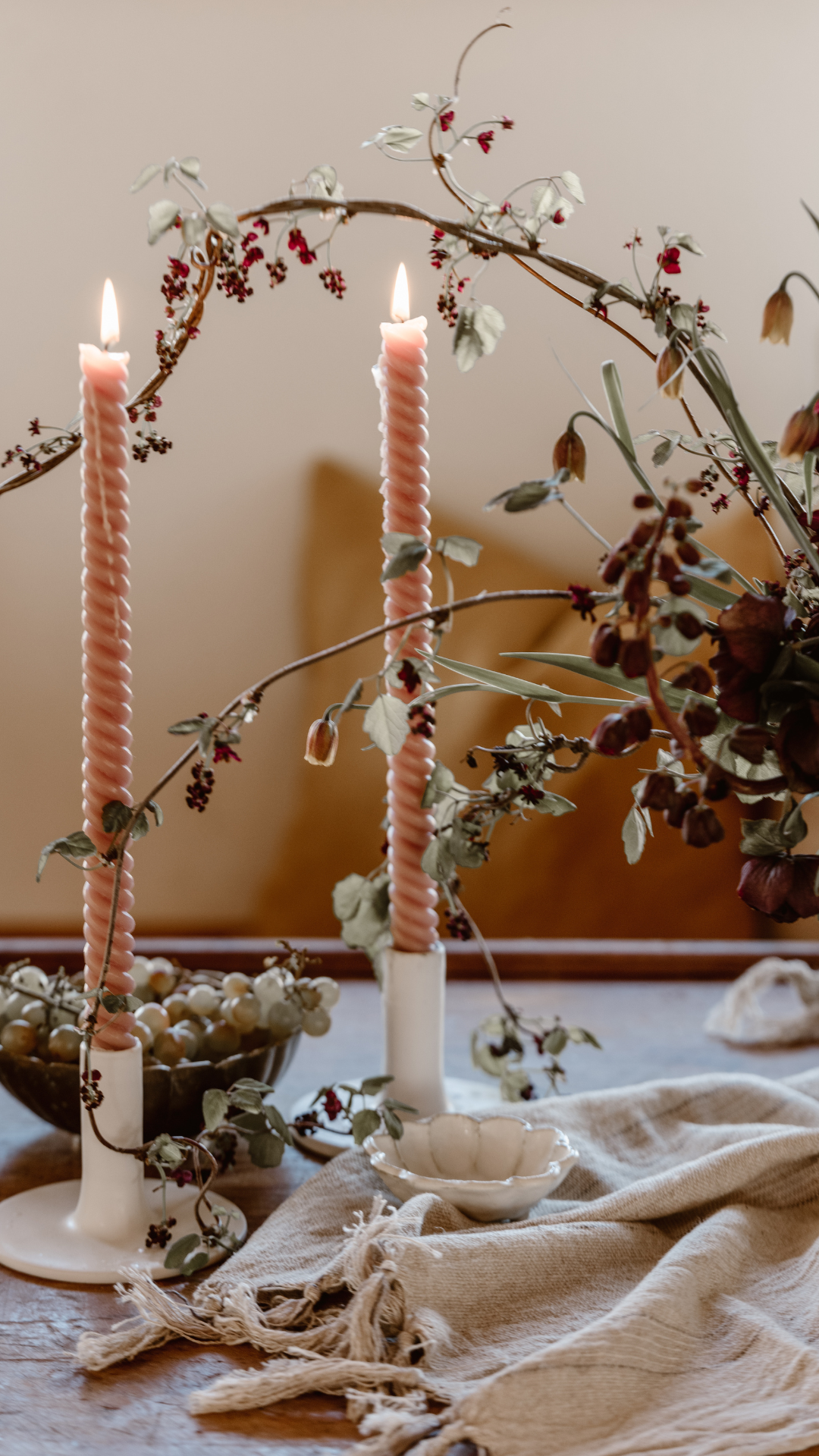 Decorative table setting with two pink twisted candles on white candle holders, dried flowers and foliage, a bowl of white berries, and a beige cloth on a wooden table.