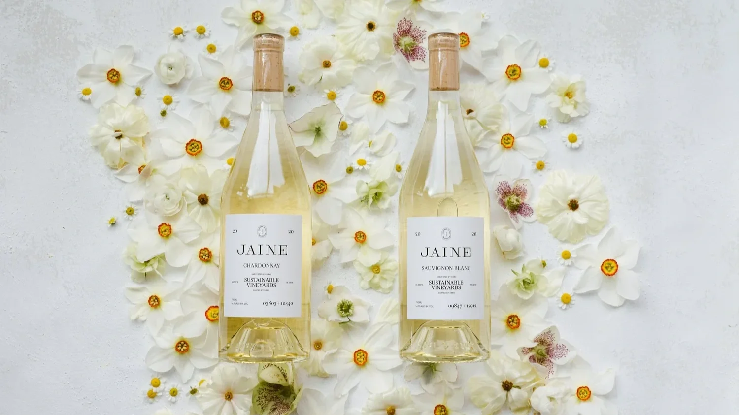 Two bottles of Jaine wine, one Chardonnay and one Sauvignon Blanc, on a white surface surrounded by white and cream-colored flowers.