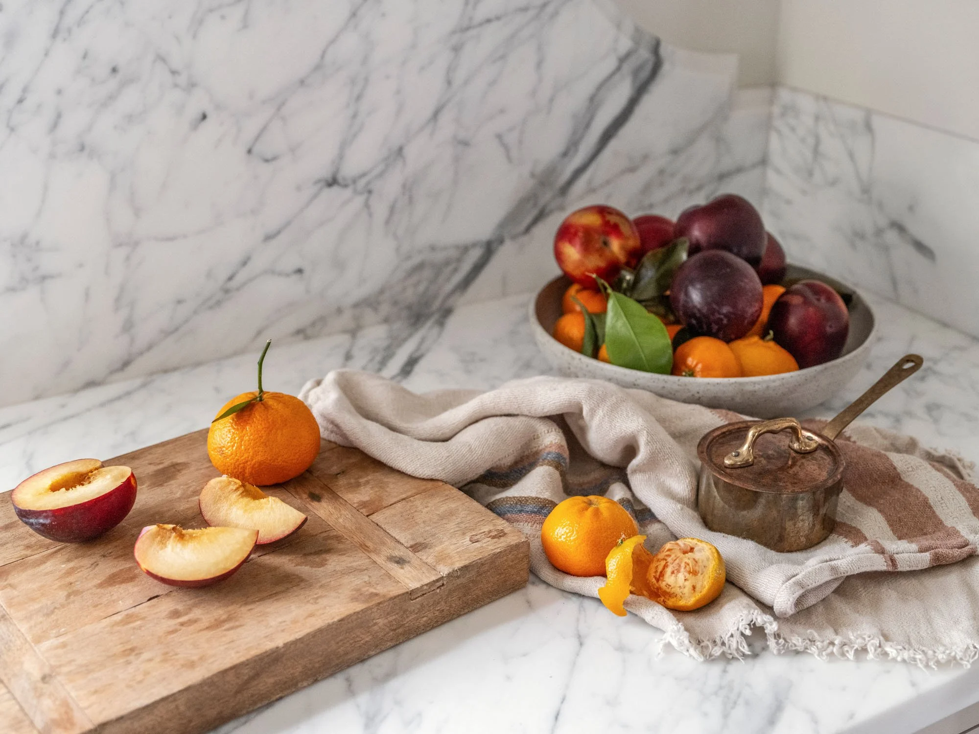 A marble kitchen countertop with a wooden cutting board holding sliced peaches and an uncut tangerine. A bowl of plums and clementines with green leaves is in the background. A beige cloth and a small copper pot are also on the counter.