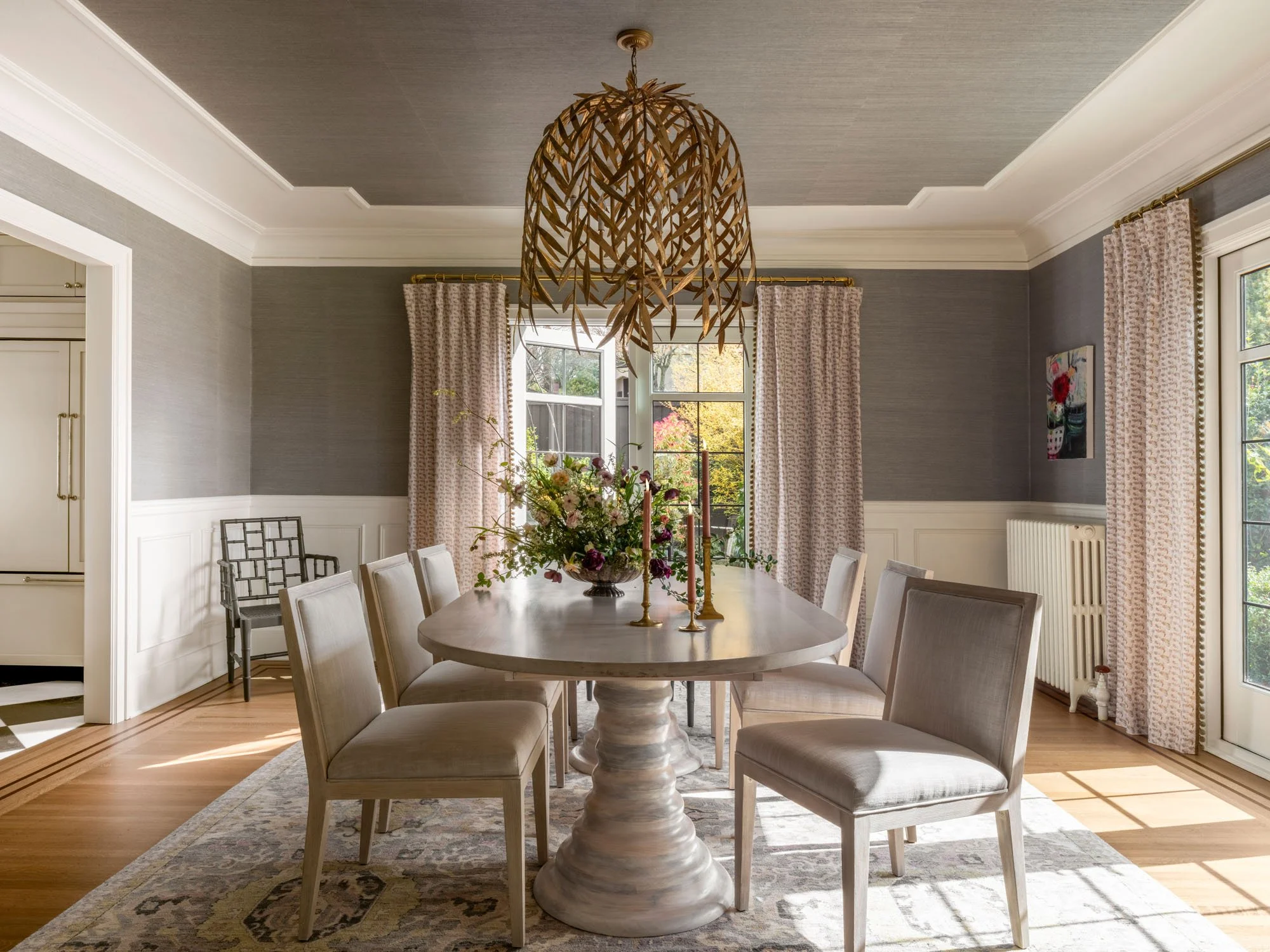 A dining room with a round table, six upholstered chairs, a floral centerpiece, and gold candlesticks. The room has gray walls, white trim, large windows with patterned curtains, and a decorative ceiling light fixture.