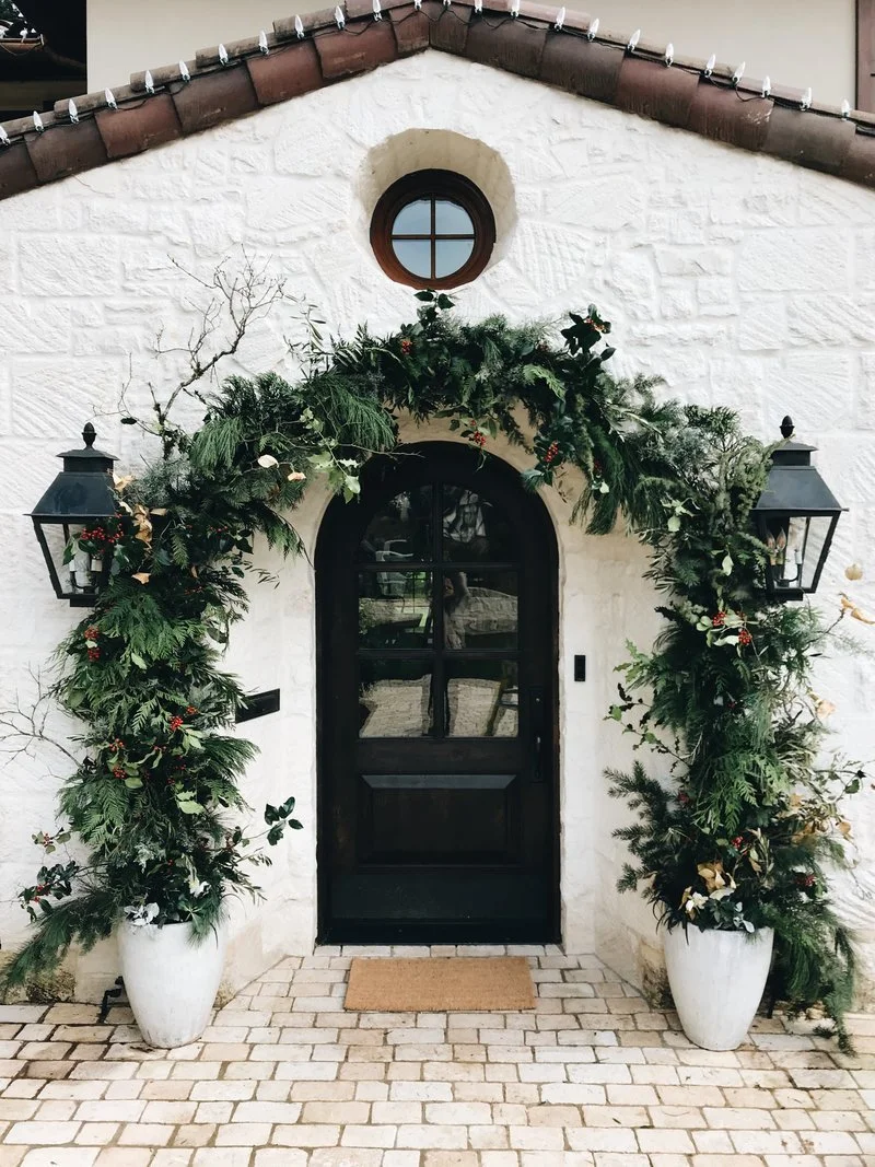 Entrance door decorated with greenery and holiday decorations, flanked by two large white planters with plants and candles, on a brick walkway, with two black outdoor lanterns on either side of the door.