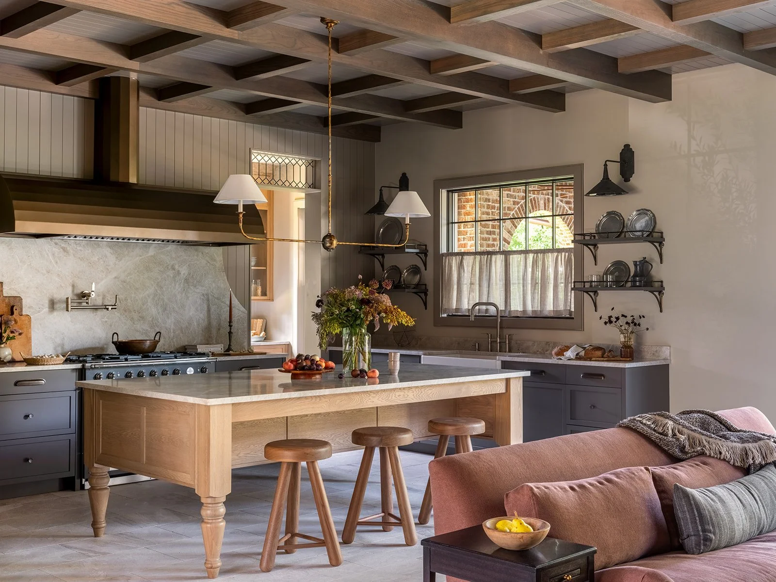 A cozy kitchen with a central kitchen island, gray cabinets, open shelves with plates, a window above the sink, and a pink sofa with a throw blanket in the adjacent living space.
