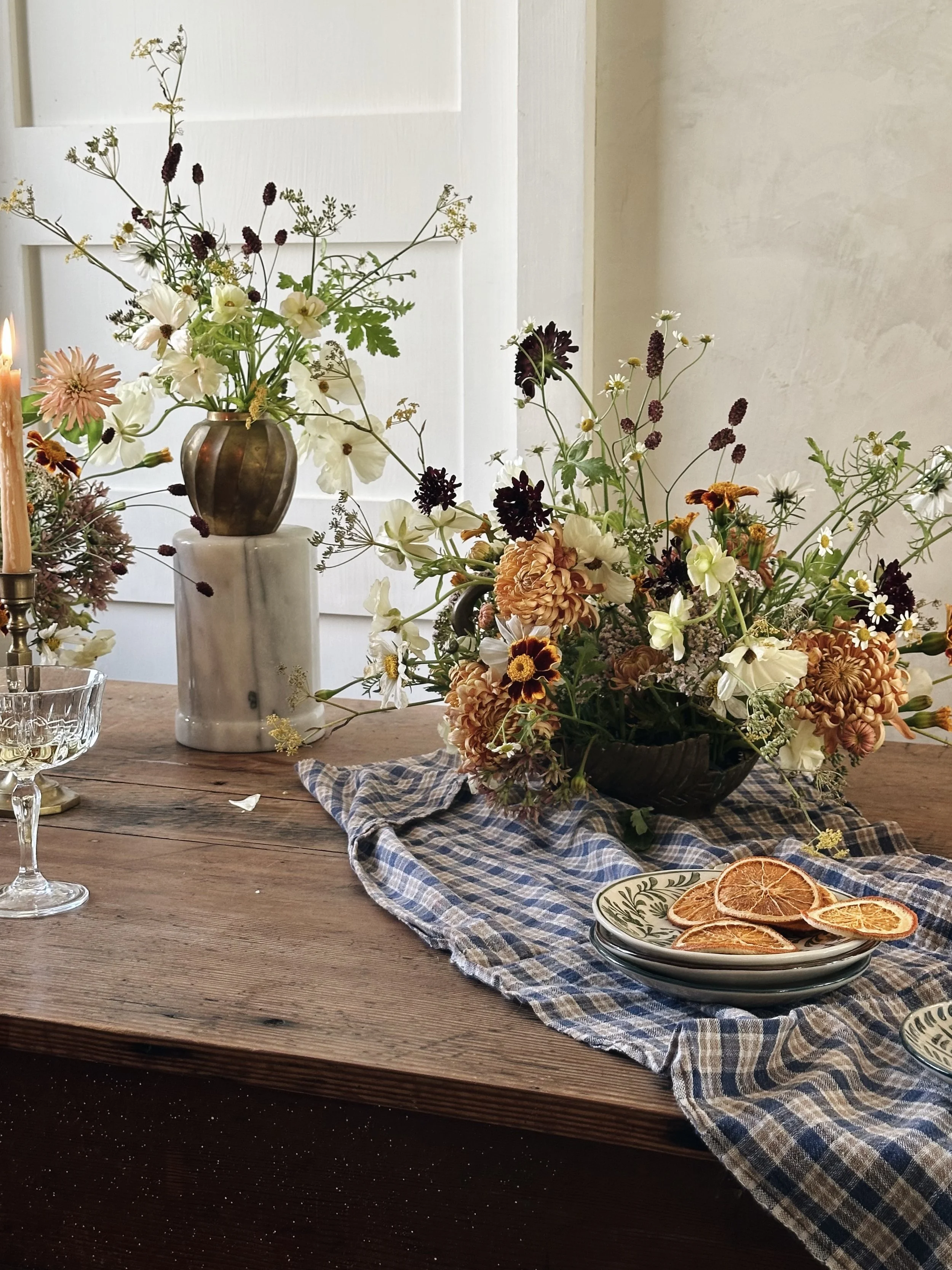 A rustic wooden table decorated with two vases of wildflowers, dried citrus slices on patterned plates, and a blue checkered table runner, with a candle and glassware nearby.