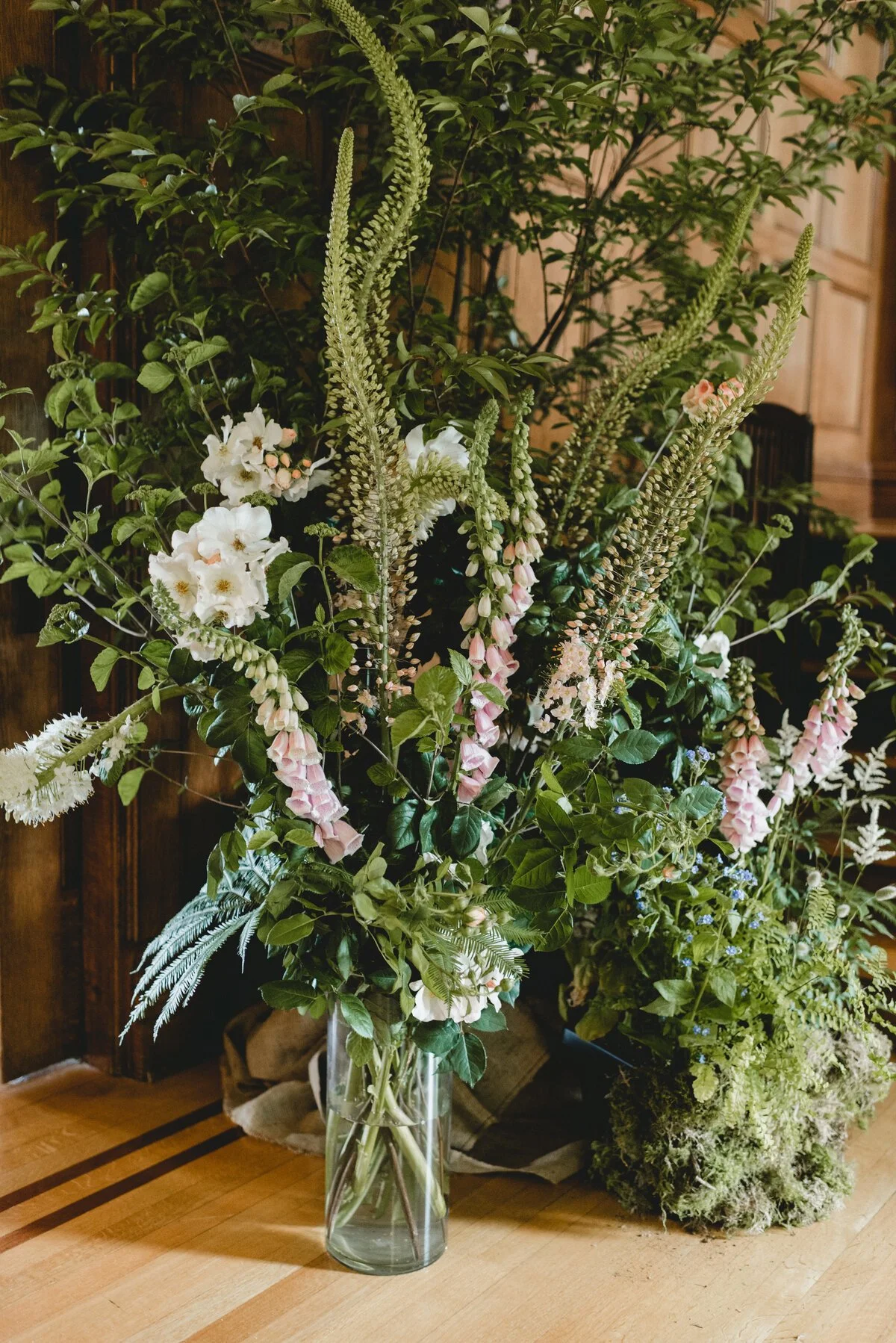 A large bouquet of mixed flowers and greenery in a tall glass vase on a wooden surface.