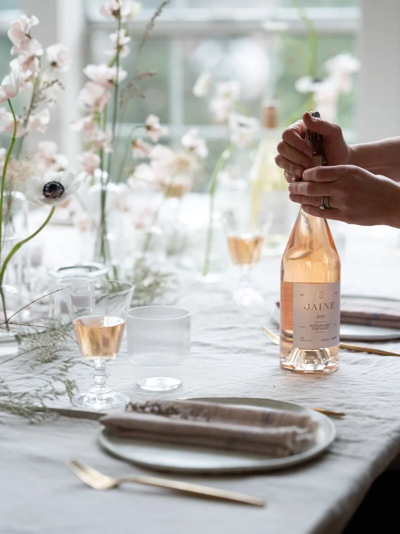 A person opening a bottle of rosé wine at a table set with glassware, plates, and cutlery, with a floral arrangement in the background.