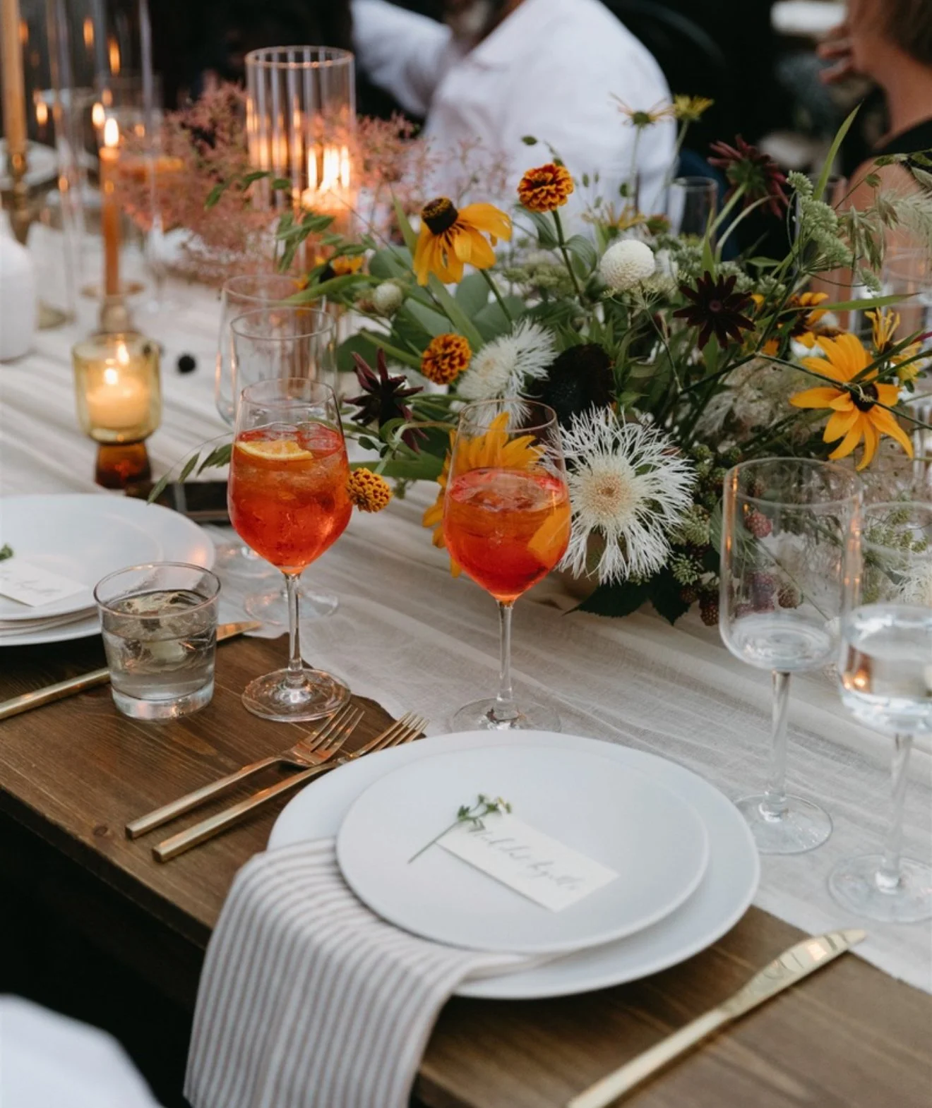 A beautifully set outdoor table with a white tablecloth, floral centerpiece, candles, and glasses of orange-colored drinks, likely during a daytime event or celebration.