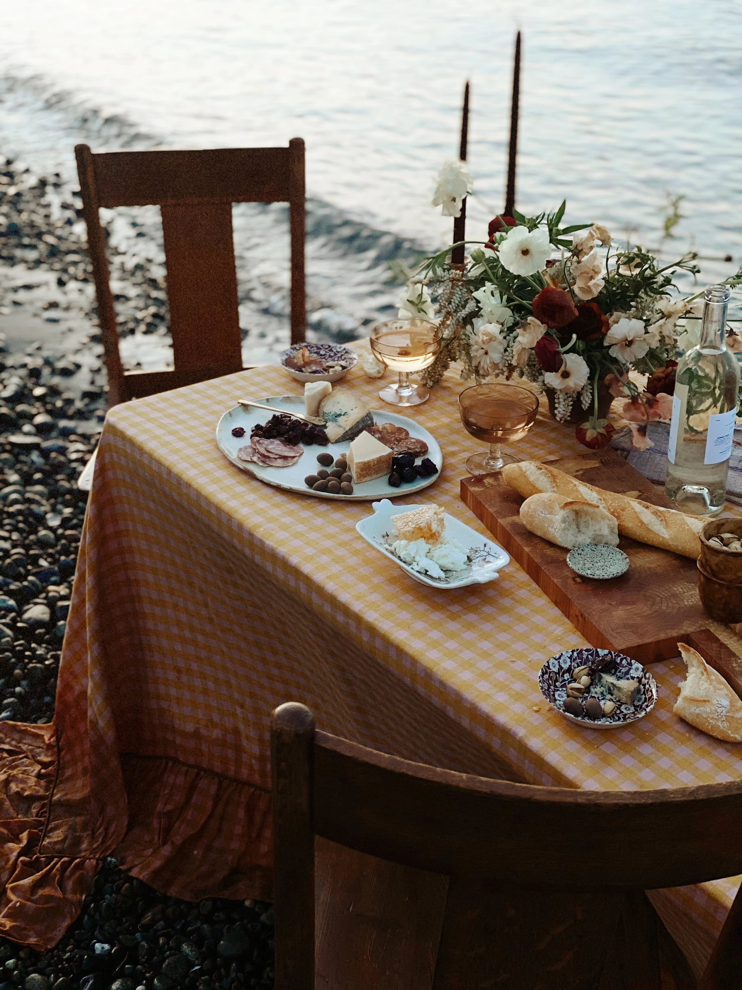 A table set for a seaside outdoor meal with a yellow checkered tablecloth, assorted cheeses, meats, bread, fruit, and a large floral centerpiece, near a rocky shoreline with water in the background.