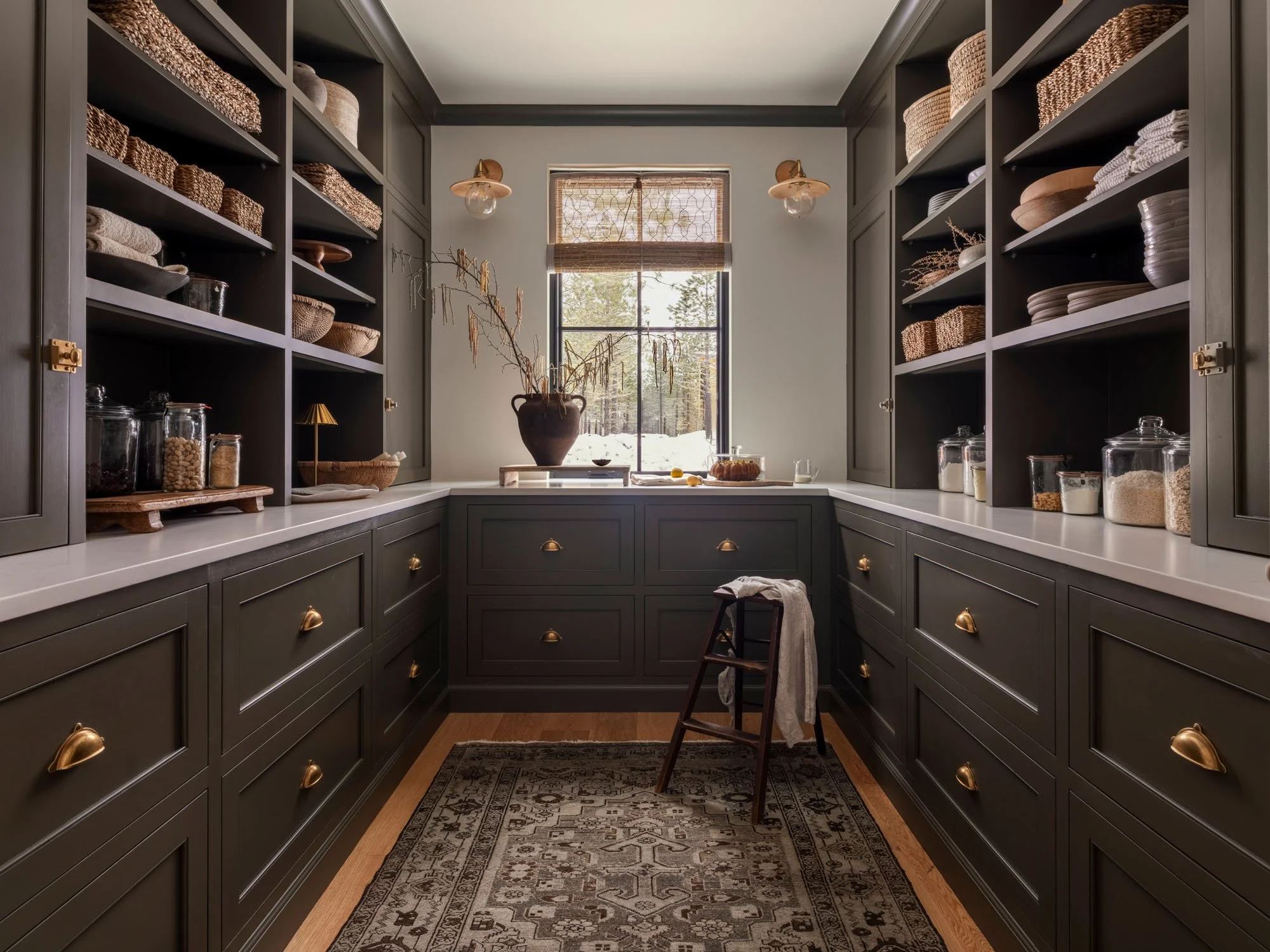 A walk-in pantry with dark gray cabinets, open shelving, and glass jars filled with food items. A window at the end provides natural light. There is a small wooden step stool and a vase with dried branches on the countertop.